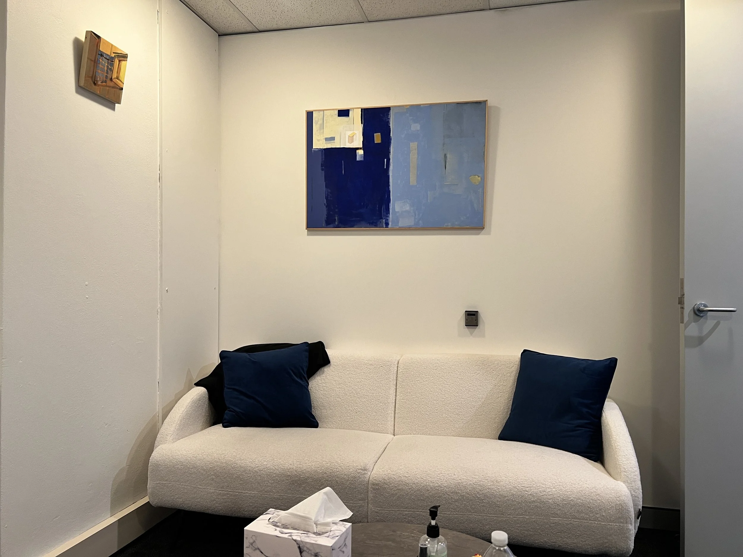 A small beige sofa with three dark blue pillows. Above the sofa, a large abstract painting with shades of blue and white is hung on the wall. To the left, a smaller framed artwork is visible. In front of the sofa, a small white table holds tissues, a hand sanitizer bottle, and a water bottle. The room has light-colored walls and a drop ceiling.