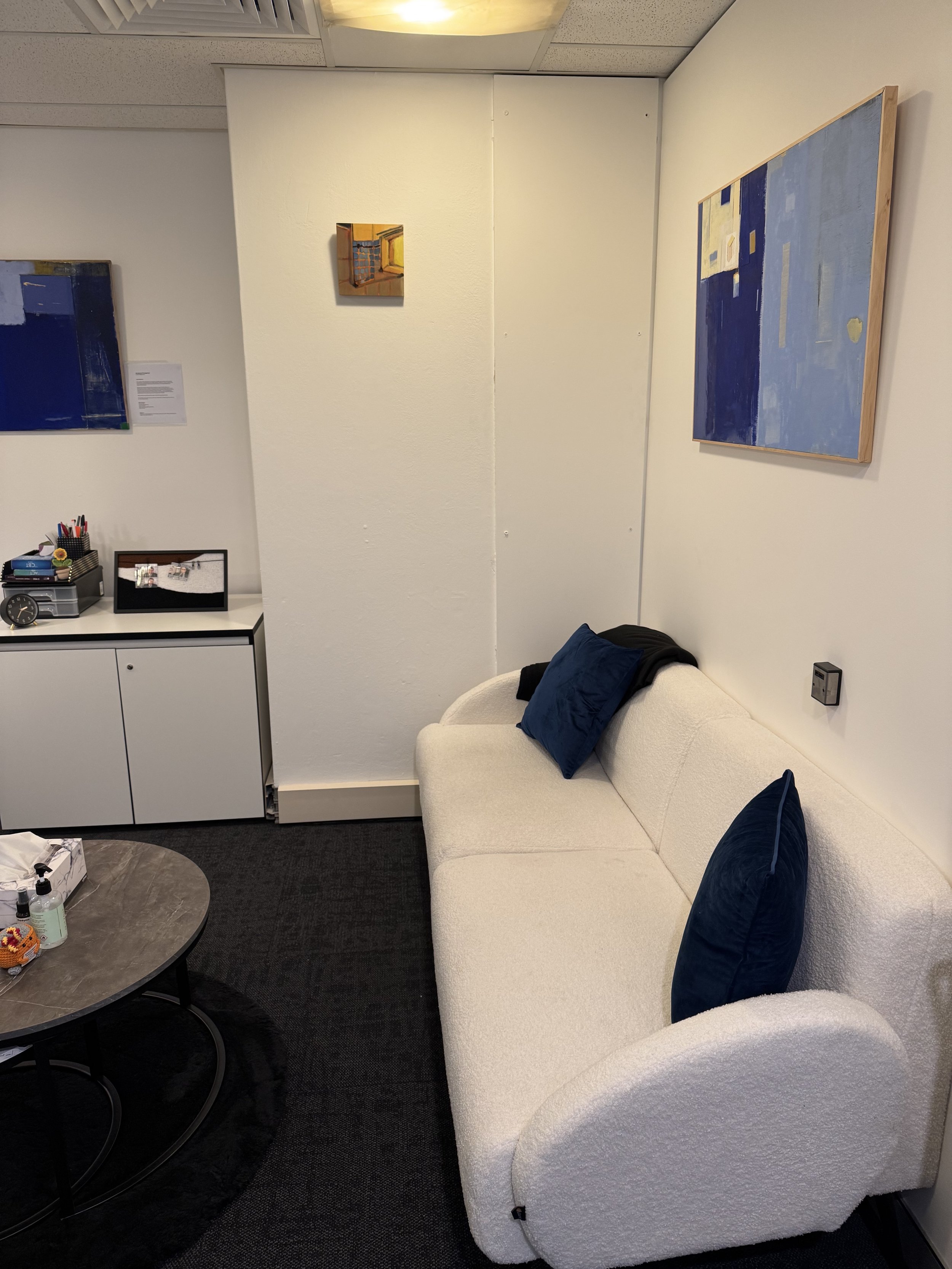 An office waiting area with a white sofa and blue pillows, a small round coffee table with bottles and a toy, a cabinet with office supplies, a television, and two paintings on the walls.