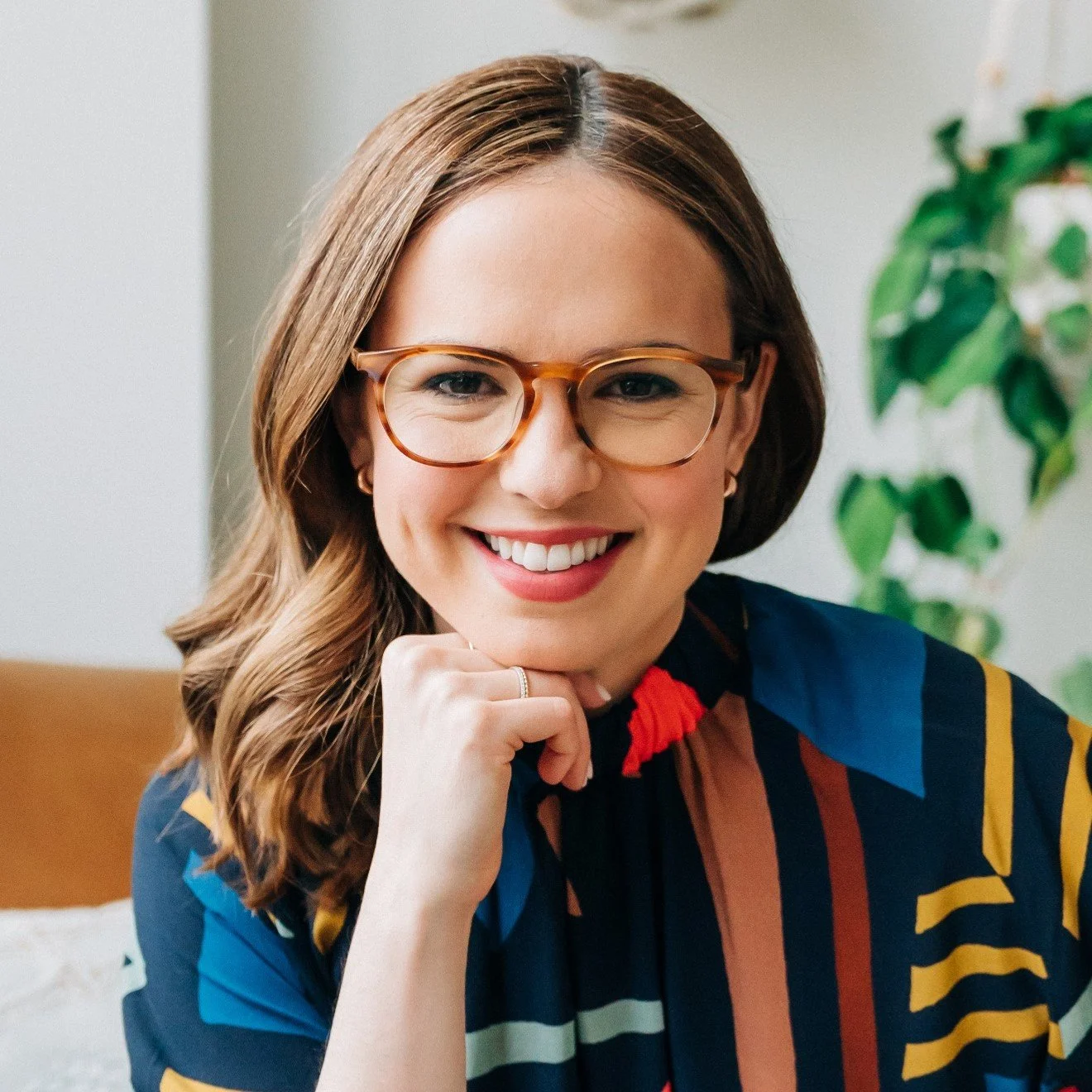 A smiling woman with brown hair, glasses, and a striped multicolored shirt, resting her chin on her hand, indoors with a plant in the background.