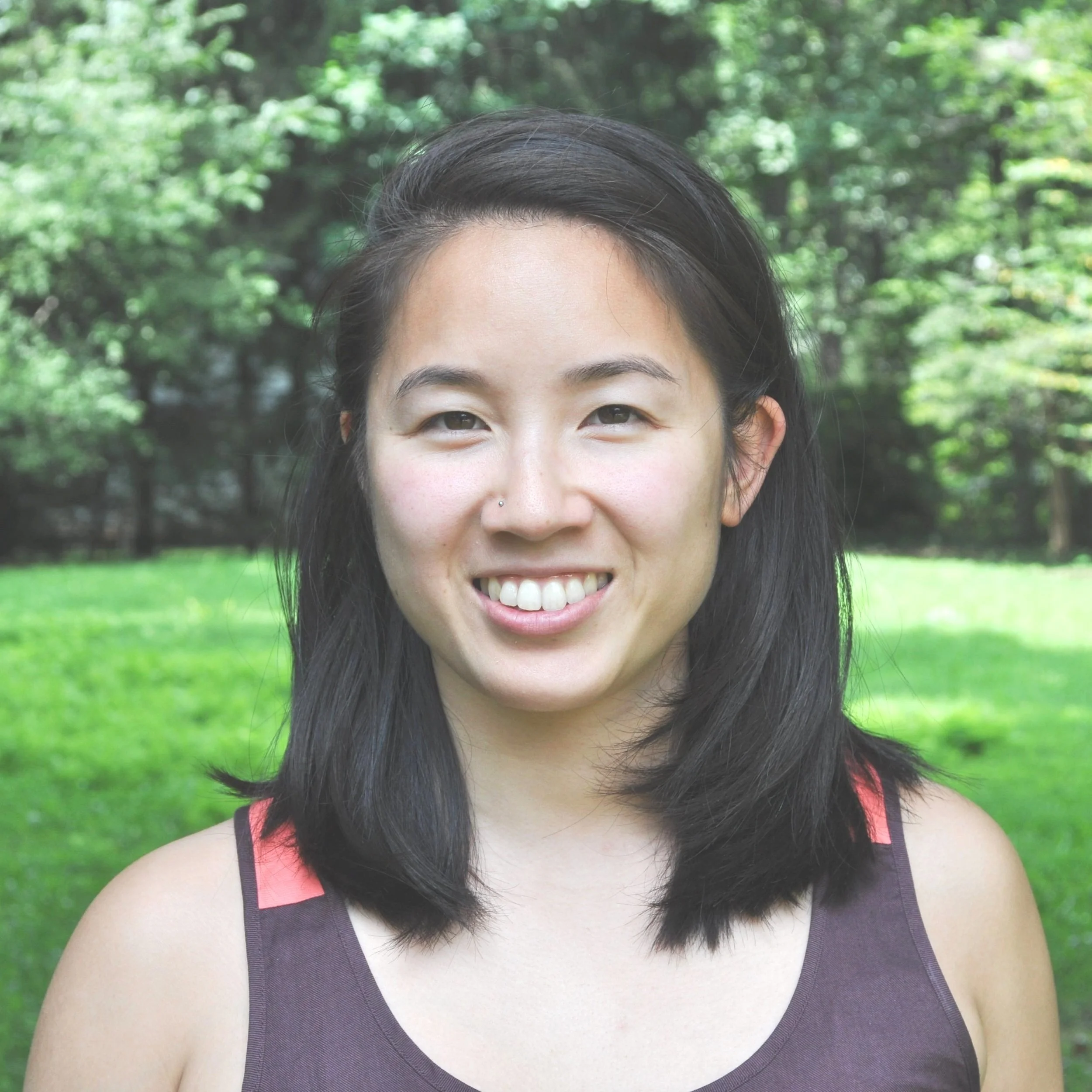 A woman with shoulder-length black hair and fair skin, smiling with teeth showing, wearing a sleeveless dark top with pink accents along the shoulders, standing outdoors in a lush green park with trees in the background.