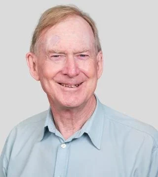 A smiling elderly man with light skin, short light hair, wearing a light blue collared shirt.