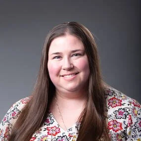 A young woman with long brown hair, smiling, wearing a white floral patterned top, against a gray background.