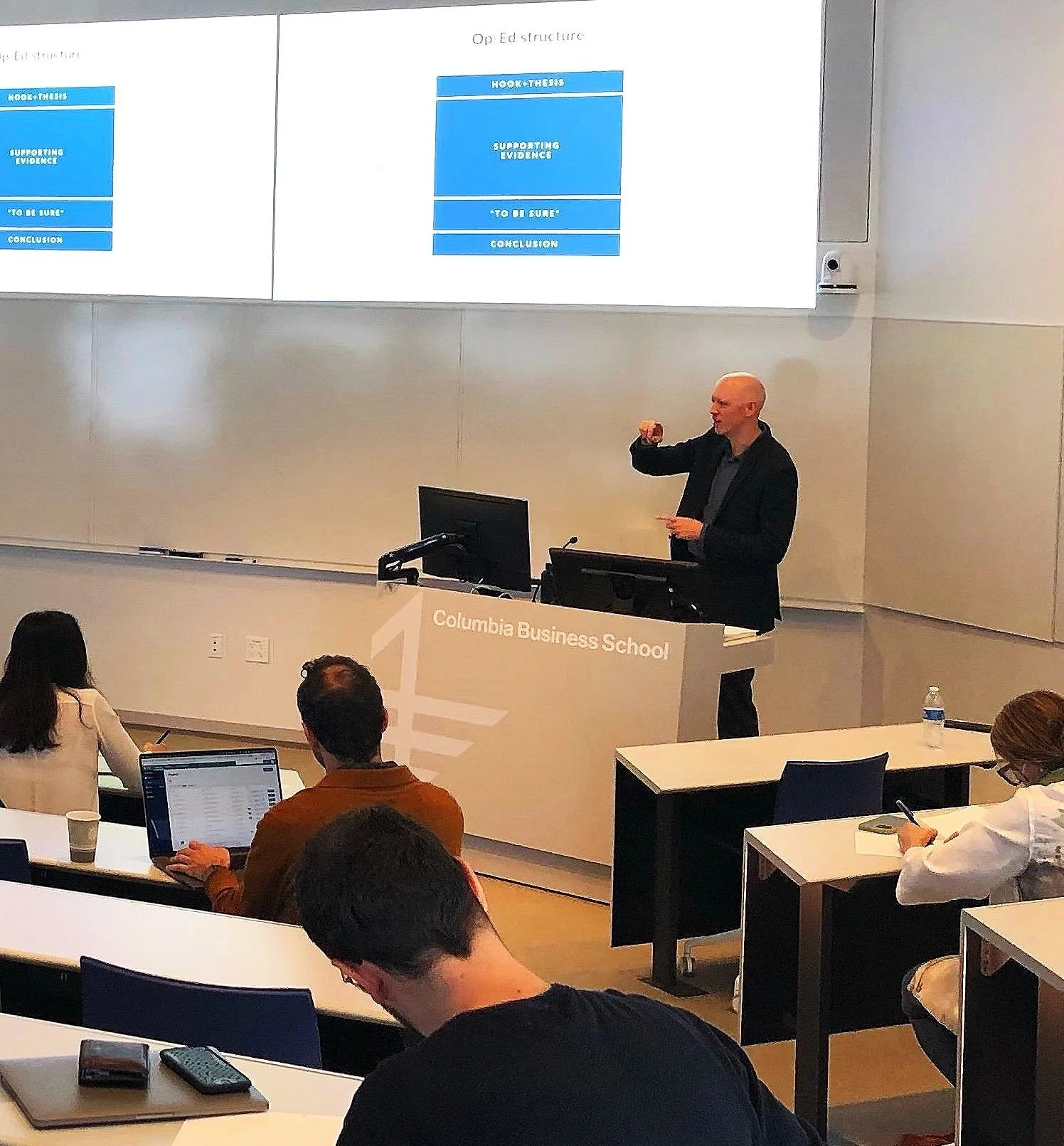 A man giving a presentation in a classroom at Columbia Business School, with slides projected on a screen behind him. Several students are seated, some using laptops and taking notes.