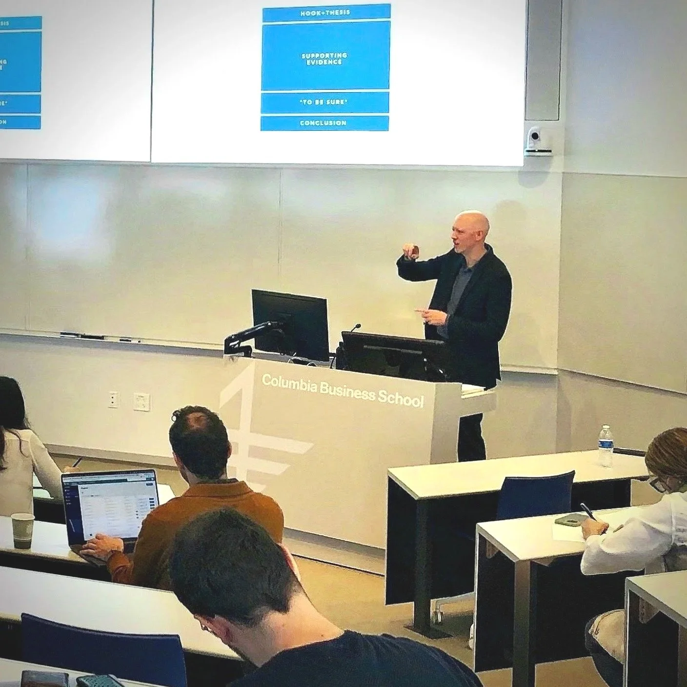 Paul Jensen delivering a training to faculty at Columbia Business School, with a large screen displaying a slide titled "Hook & Thesis". Audience members are seated, some taking notes or using laptops.