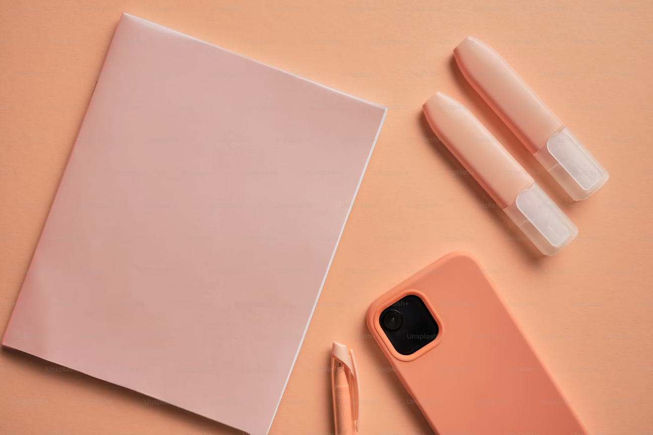 Flat lay of a peach-colored smartphone, two peach-colored lip glosses, a peach-colored notebook, a peach-colored pen, on a peach background.