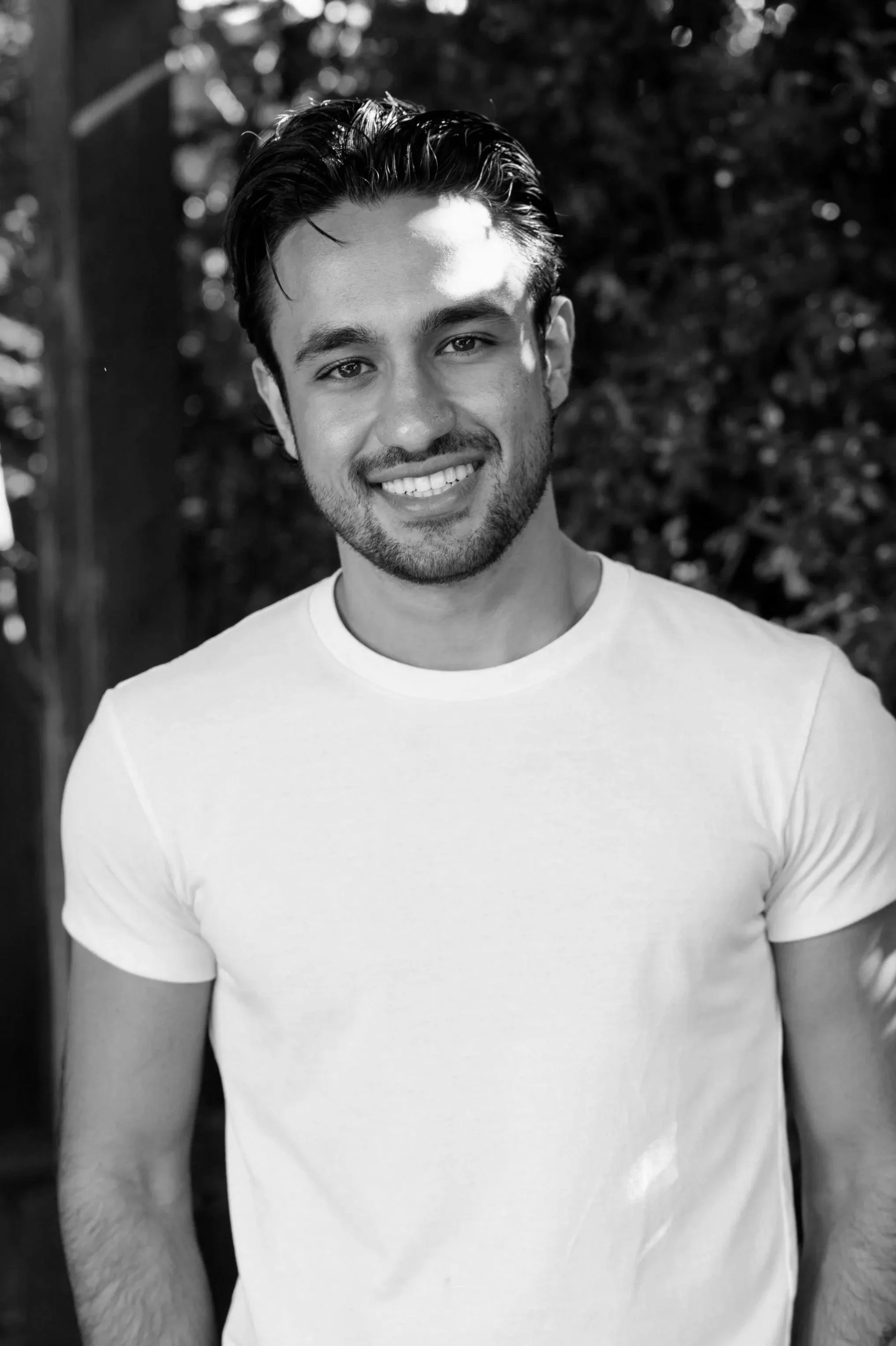 A young man smiling outdoors in a black and white photo, wearing a plain t-shirt.