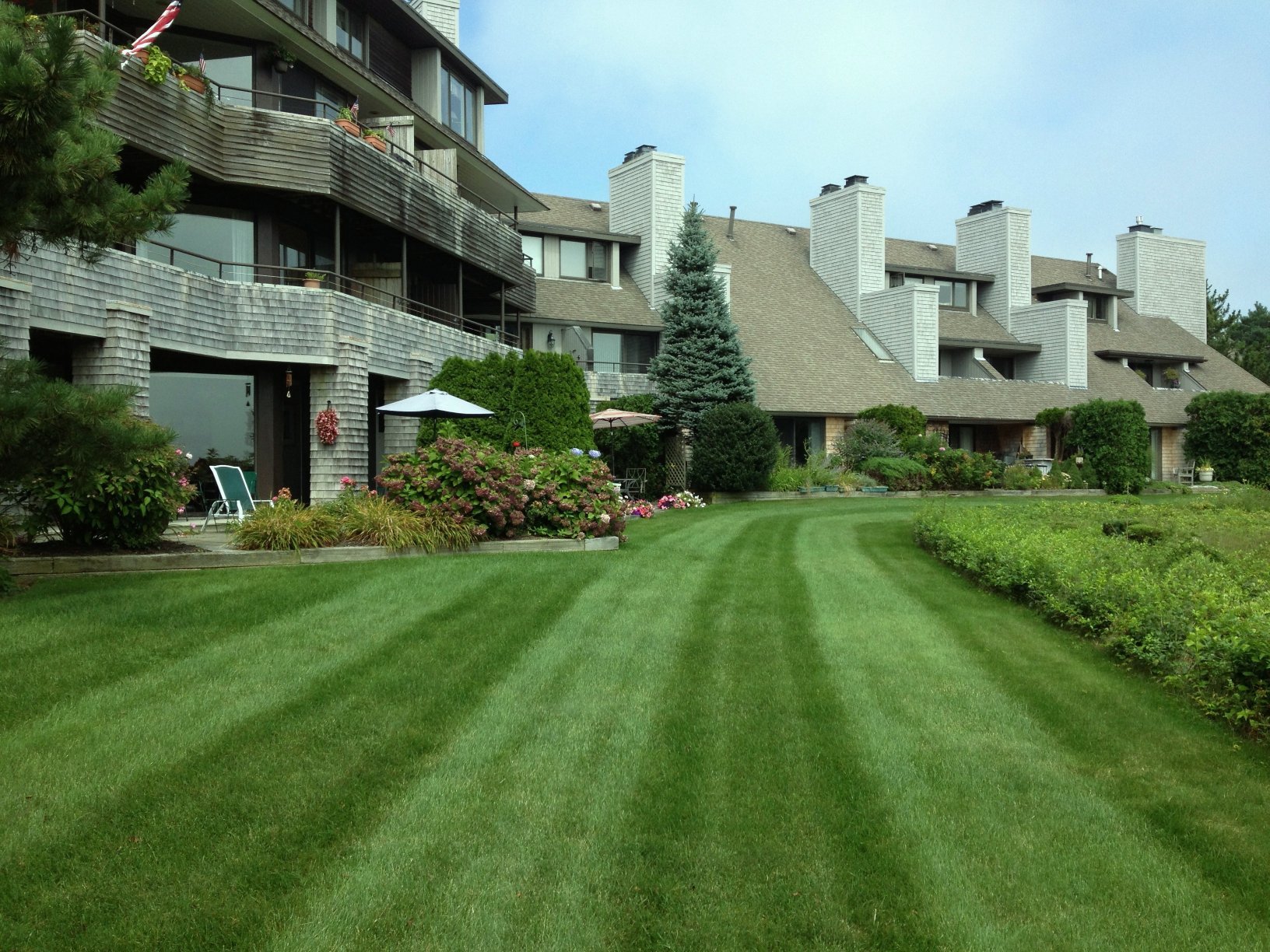 A well-maintained grassy lawn in front of a multi-story residential complex with balconies, trees, and garden furniture.