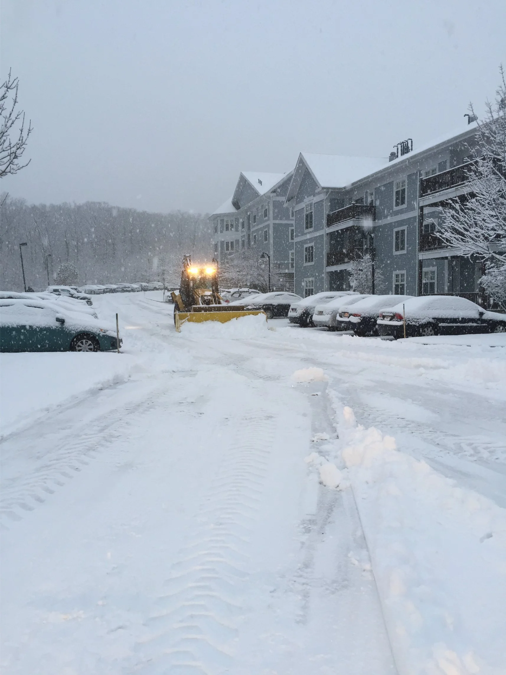 A snow-covered parking lot with a snowplow clearing the driveway during a snowfall, nearby apartment buildings, and parked cars.