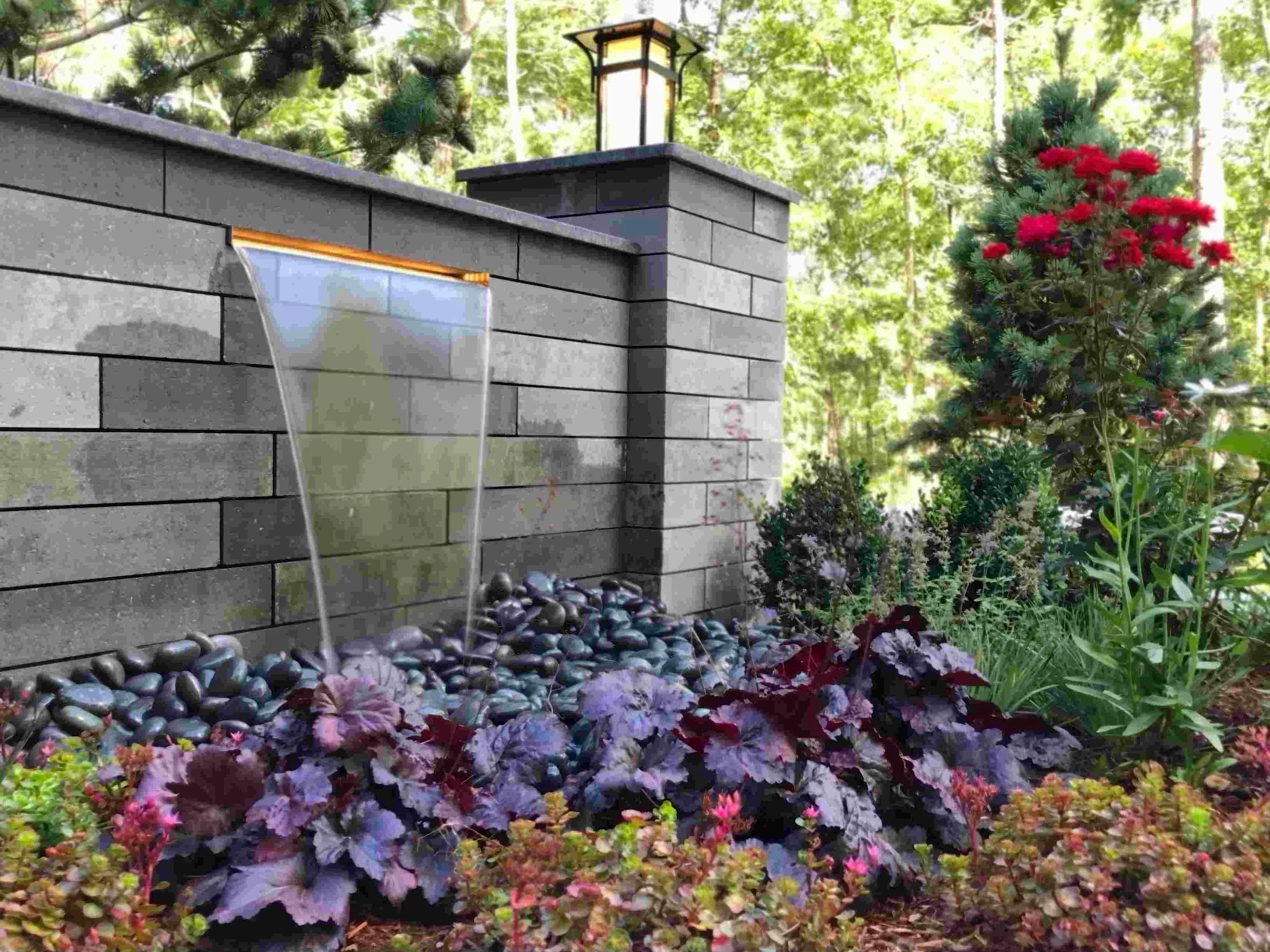 A landscaped garden with a stone wall featuring water flowing from a modern water feature, surrounded by various green plants and purple-leaved foliage, with a lantern-style light fixture on top of the wall.