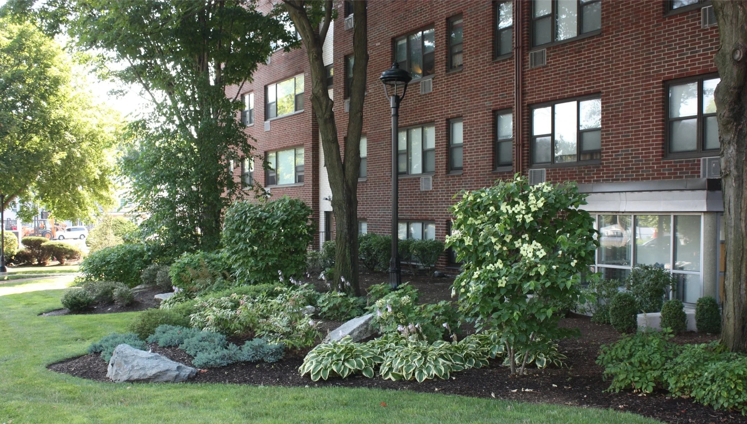 Apartment building with landscaped garden and trees in the foreground, brick exterior, and multiple windows.