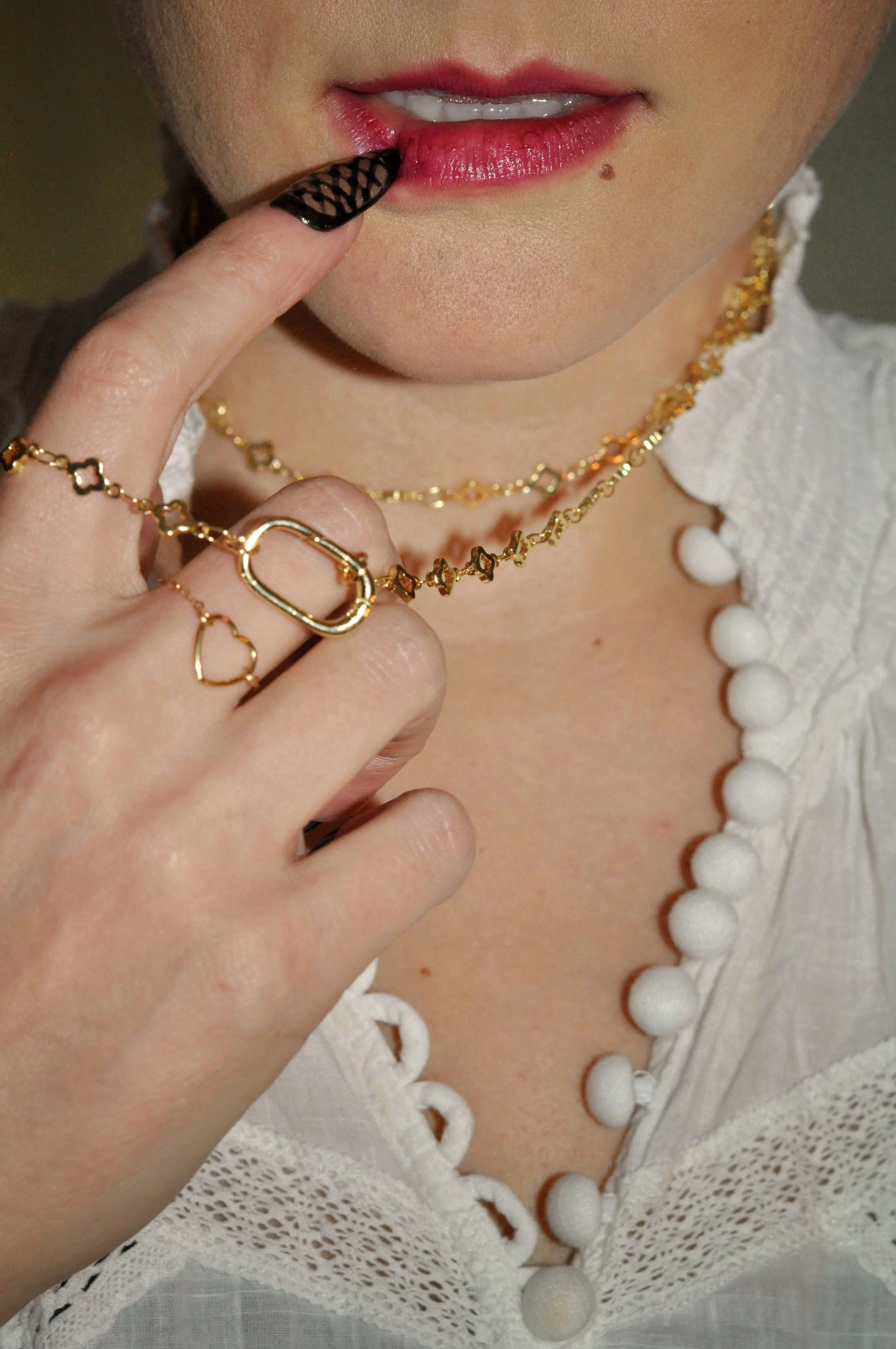 Close-up of a woman's lips, finger with black patterned nail, and jewelry including gold rings and layered necklaces, wearing a white top with decorative buttons.