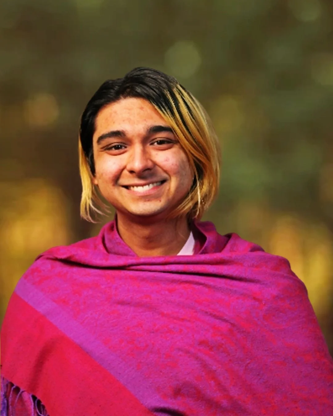 A person with brown skin smiling, wearing a pink and purple shawl, with a blurred outdoor background.