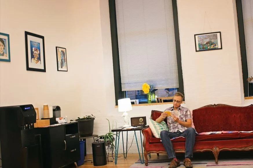 A transmasculine person sitting on a red vintage sofa, reading a book, with a window and decorative windowsill behind them, in a cozy living room with framed pictures on the wall, a  tea station, and a small table with a lamp and a radio.