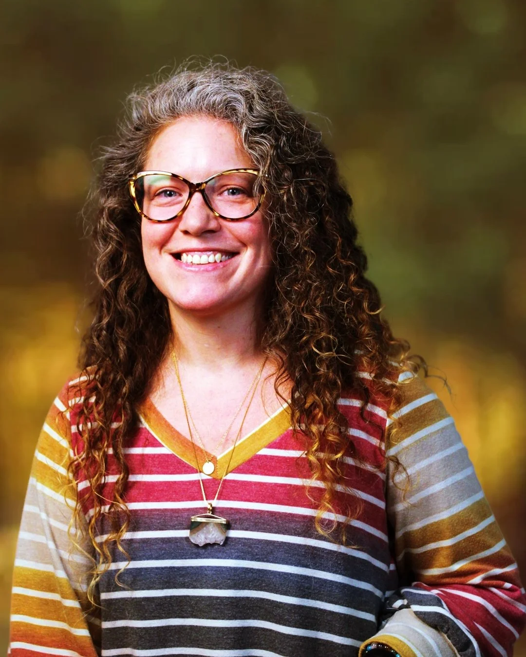 A light-skinned smiling woman with curly hair, wearing glasses, layered necklaces, and a striped shirt, outdoors with a blurred green and yellow background.