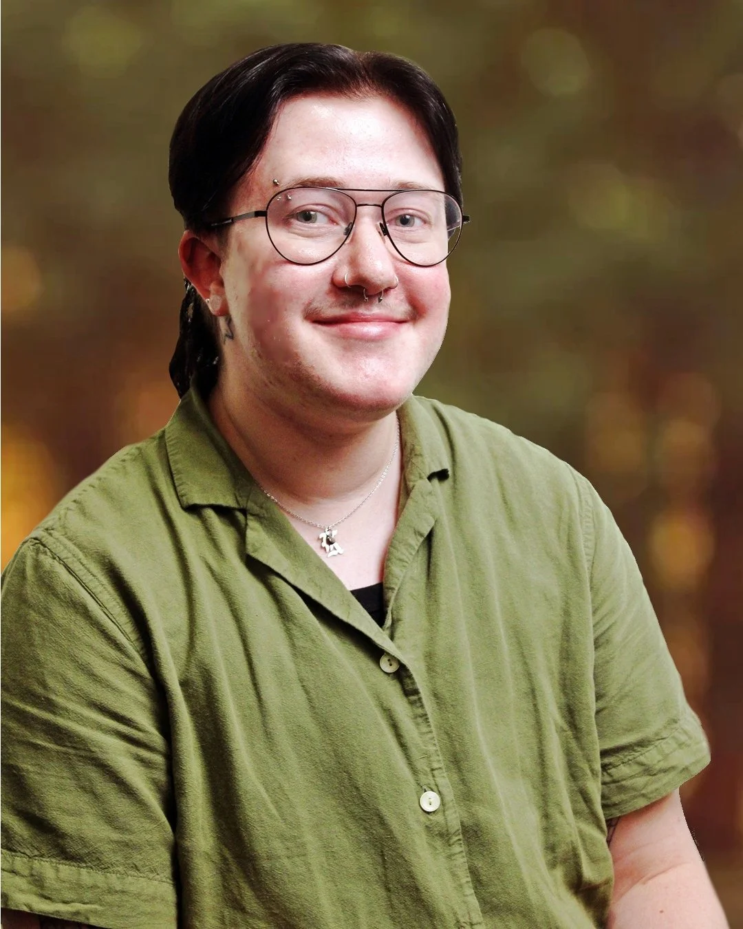A light-skinned person and short dark hair, glasses, a septum piercing, smiling and wearing a green button-up shirt against a blurred outdoor background.