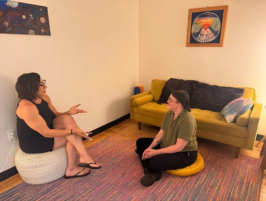Two people engaging in a conversation in an office. One is sitting on a cushion near the wall, gesturing with her hands, while the other is sitting cross-legged on a cushion on the floor facing her. There is a couch and colorful artwork on the walls.