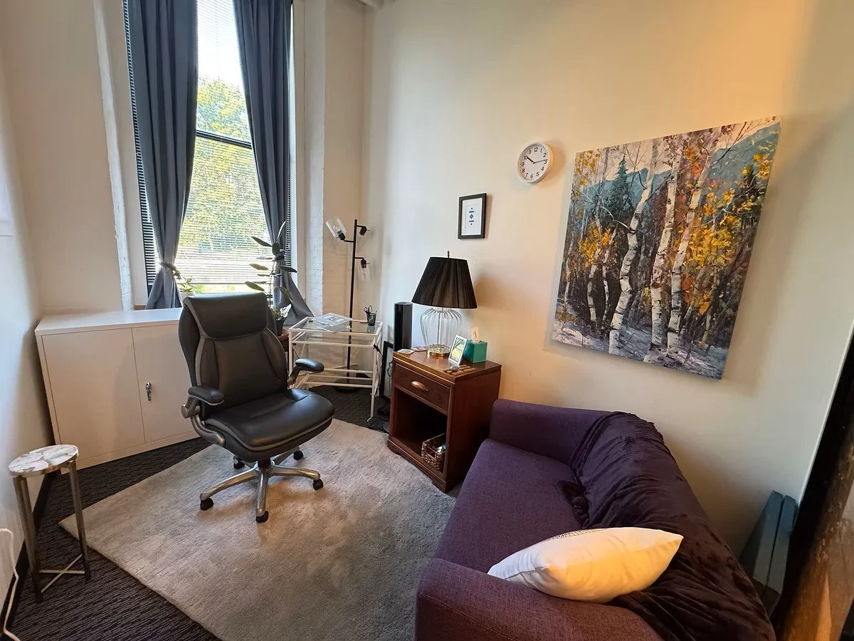 A small workspace with a black office chair in front of a window with blue curtains, a purple sofa with a white pillow, a wooden side table, a black lamp, a large landscape painting, and a clock on the wall.