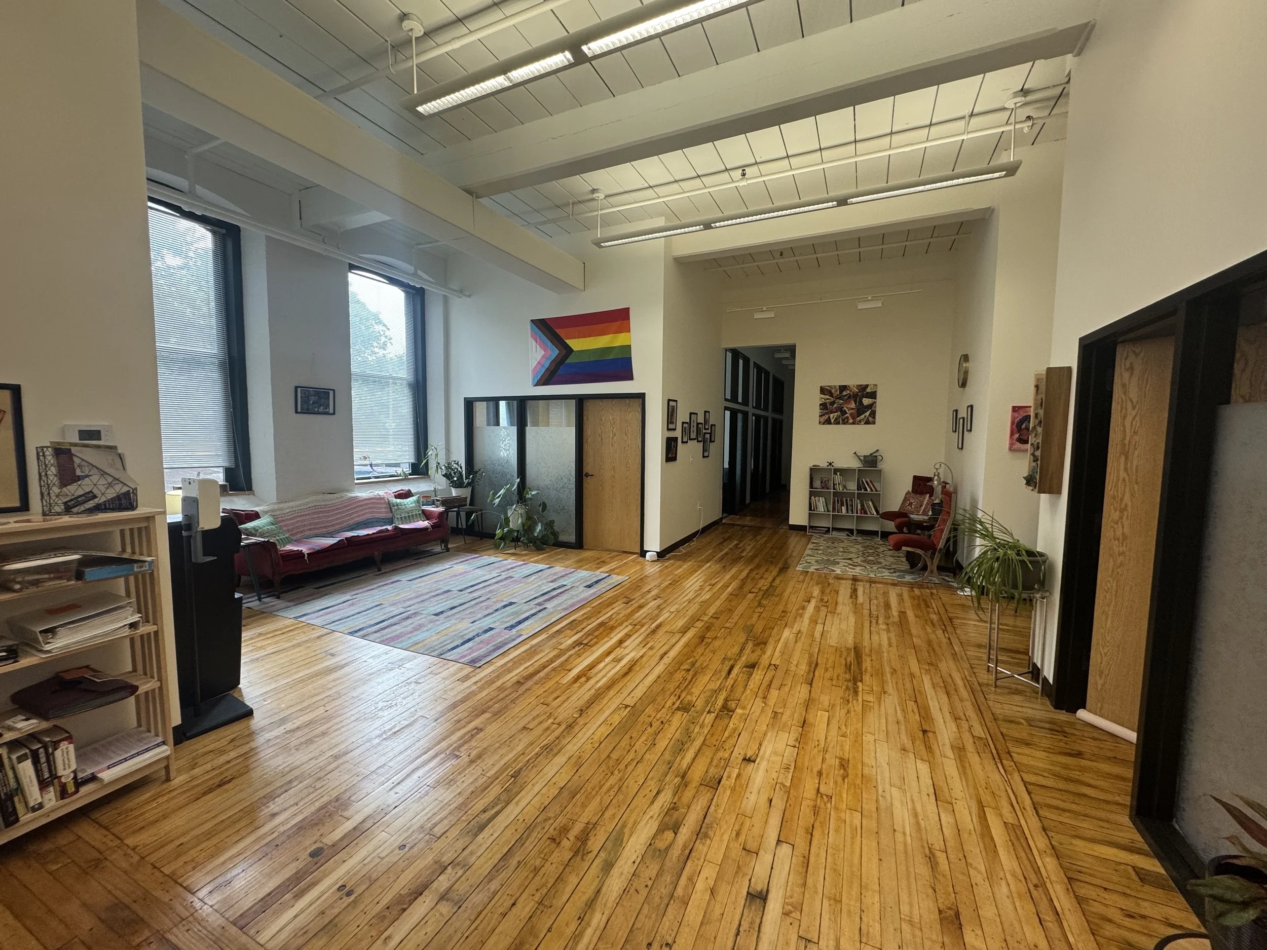 Spacious lobby with hardwood floors, large windows, and a colorful rainbow flag hanging on the wall. There is a red velvet couch, a small sitting area with a bookshelf. The room is decorated with plants and artwork by queer and trans artists.