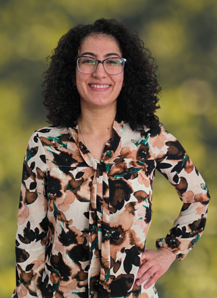 An Arab woman with Black hair in a white and brown shirt standing with her hand on one hip in front of a green background