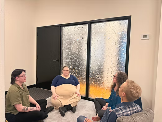 Four people sitting on the floor in a circle in front of a glass door with water droplets on it, room with beige walls and gray carpet.