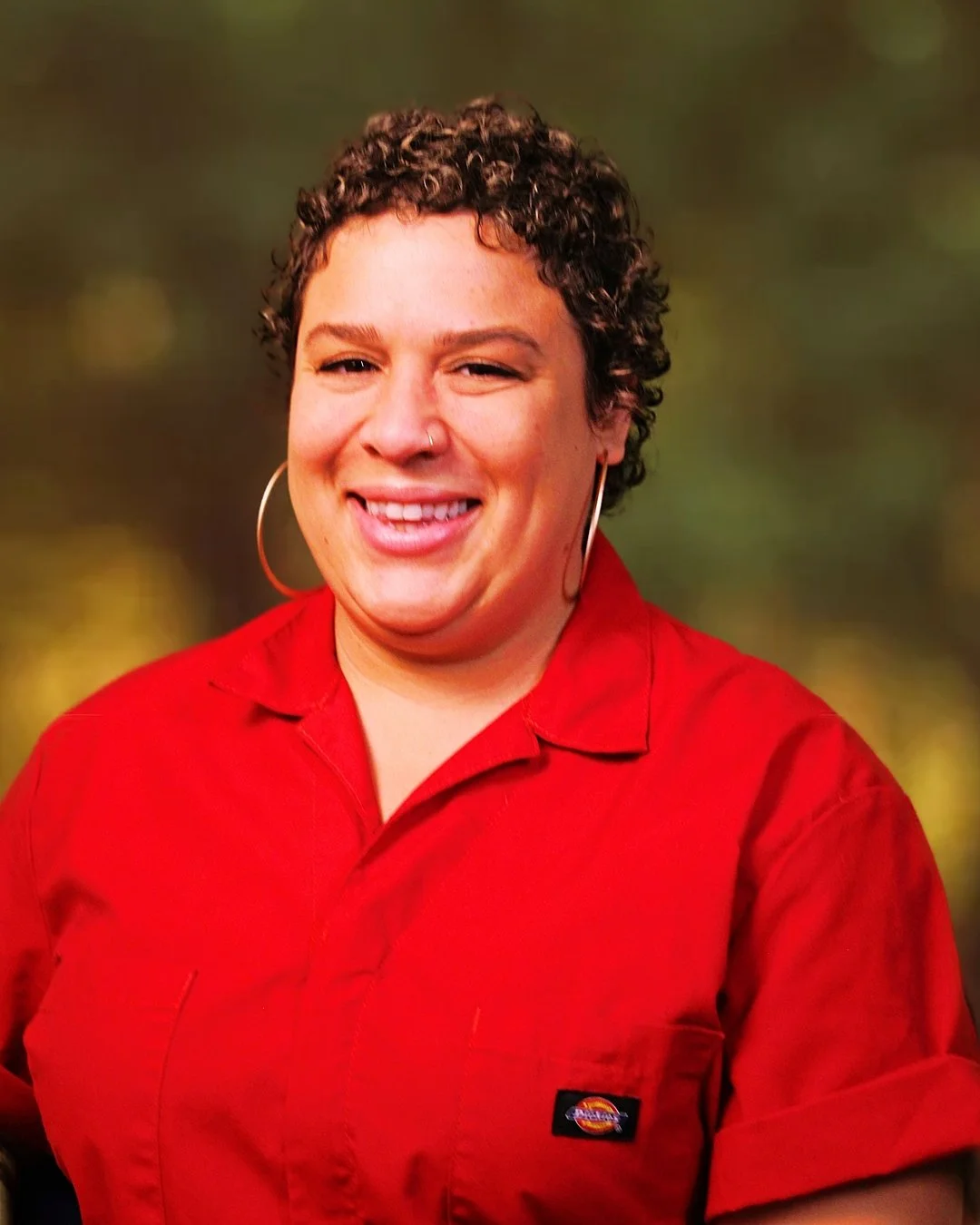 A person with brown skin and short curly hair, wearing hoop earrings and a red button-up shirt, smiling with a blurred outdoor background.