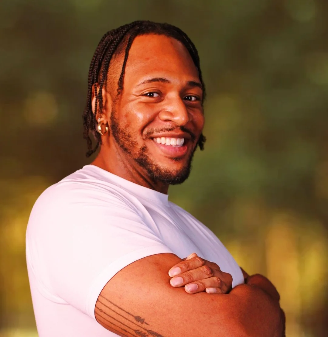 A smiling man with brown skin, braided hair, wearing a white t-shirt and earrings, standing outdoors with arms crossed against a blurred green background.