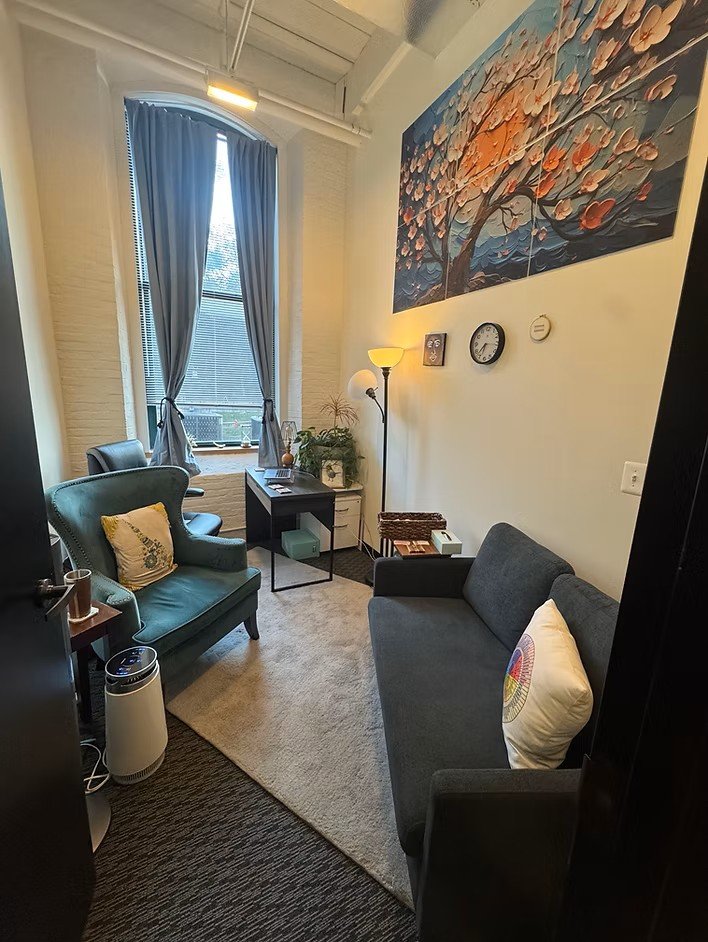 Small, cozy office space with a large window, blue curtains, a green armchair with a decorative pillow, a gray sofa with a colorful pillow, a small black desk with office supplies, a standing lamp, a tall floor lamp, and artwork of a flowering tree on the wall.