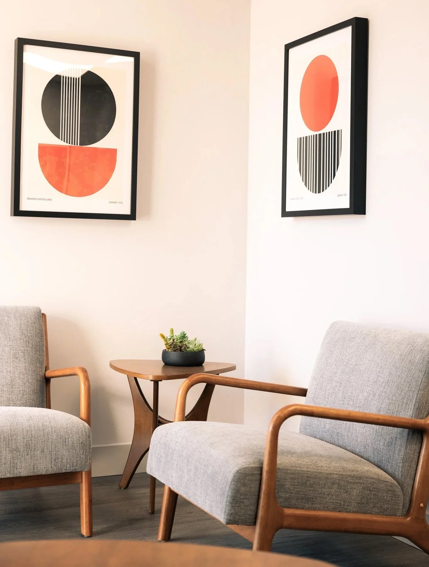 A modern living room corner with two gray armchairs with wooden arms, a small wooden side table with a plant, and two abstract framed art pieces on the white wall.