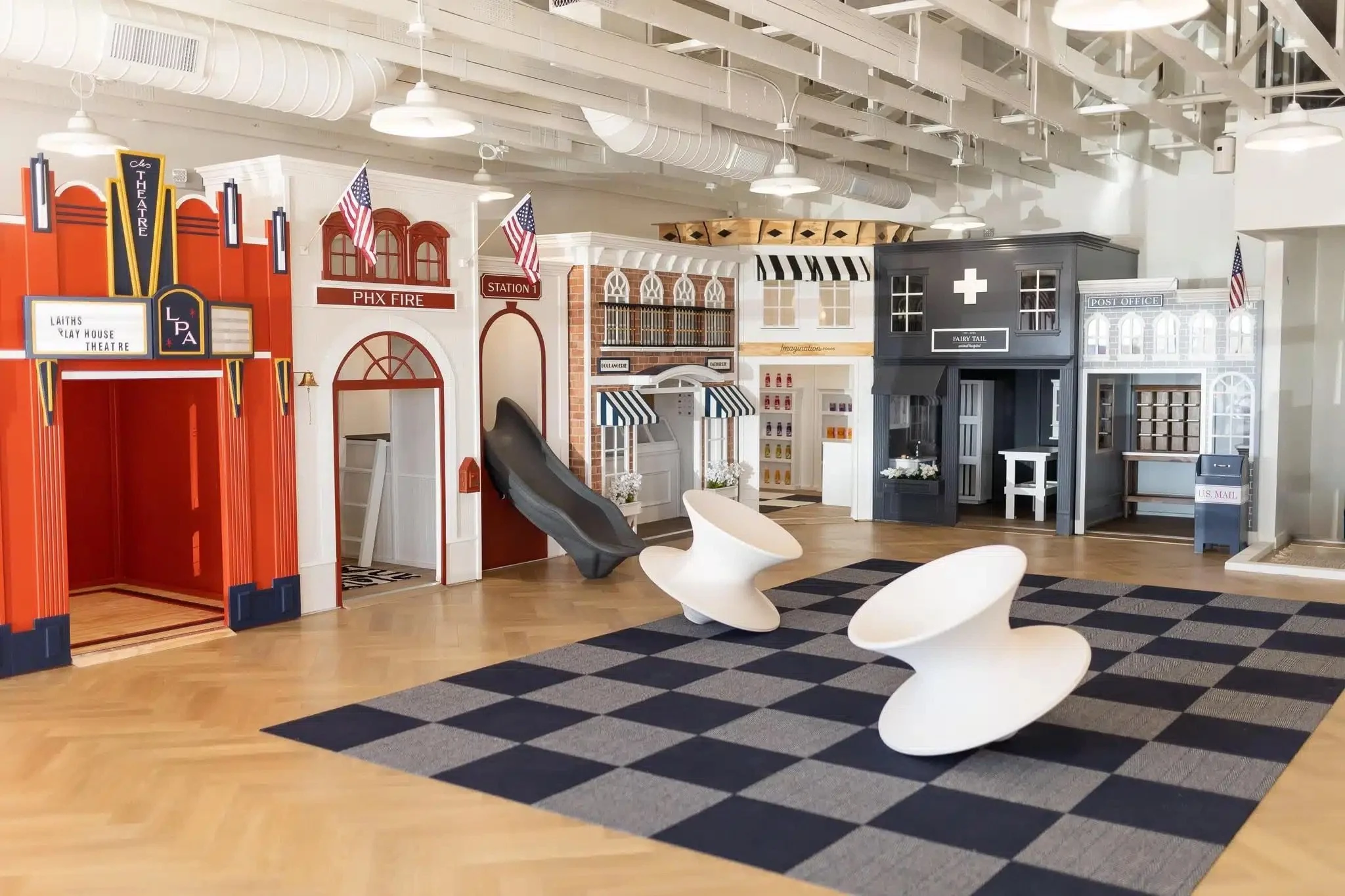 Colorful indoor play area with a small town setup, including a theater, fire station, post office, and shops, with modern white chairs and a checkered rug in the foreground.