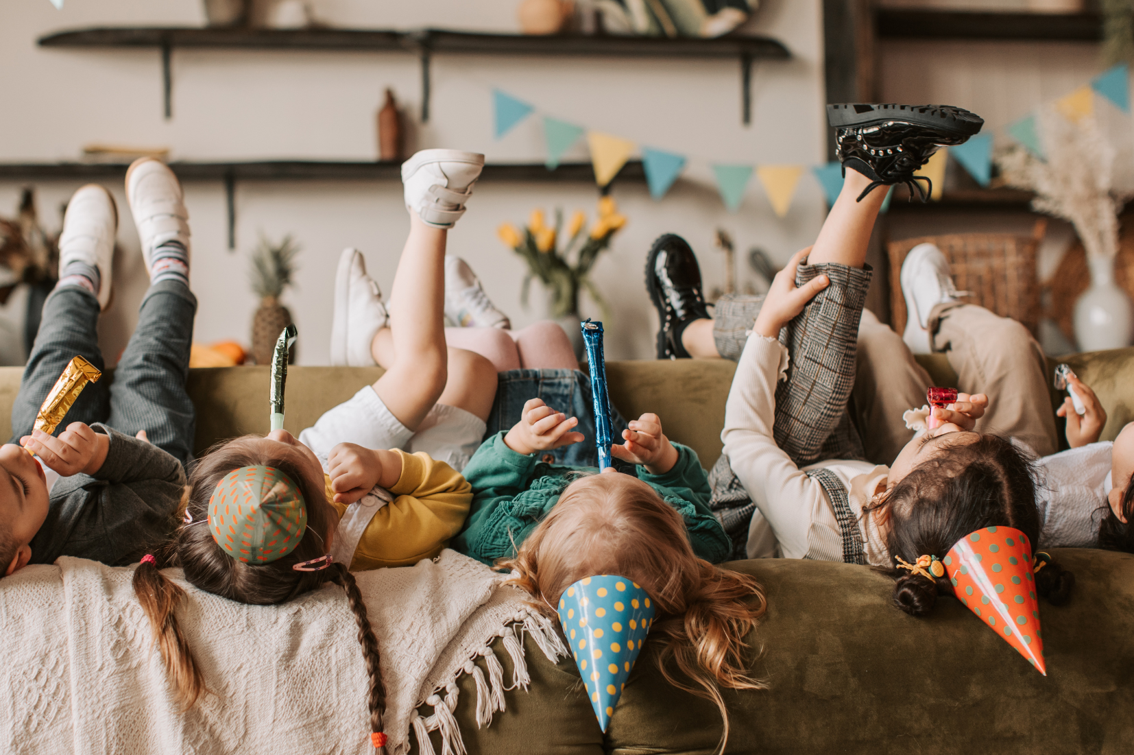 Children lying on a sofa in party hats, holding noise makers and snacks, celebrating a birthday or celebration indoors.