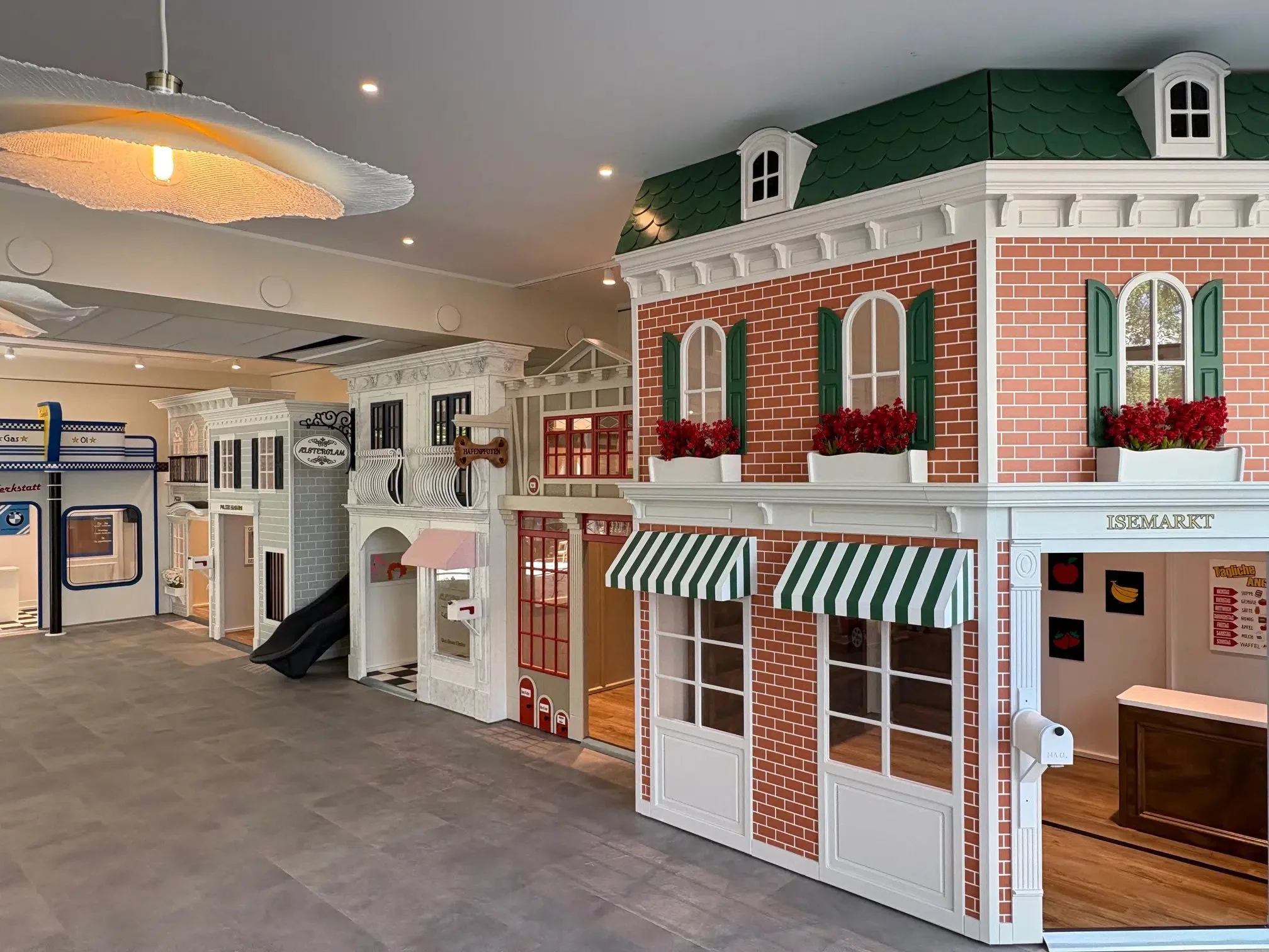 Interior of a children's play area designed to look like a miniature town with small building facades, including a brick building with flower boxes and striped awnings, and other storefronts with decorative facades.