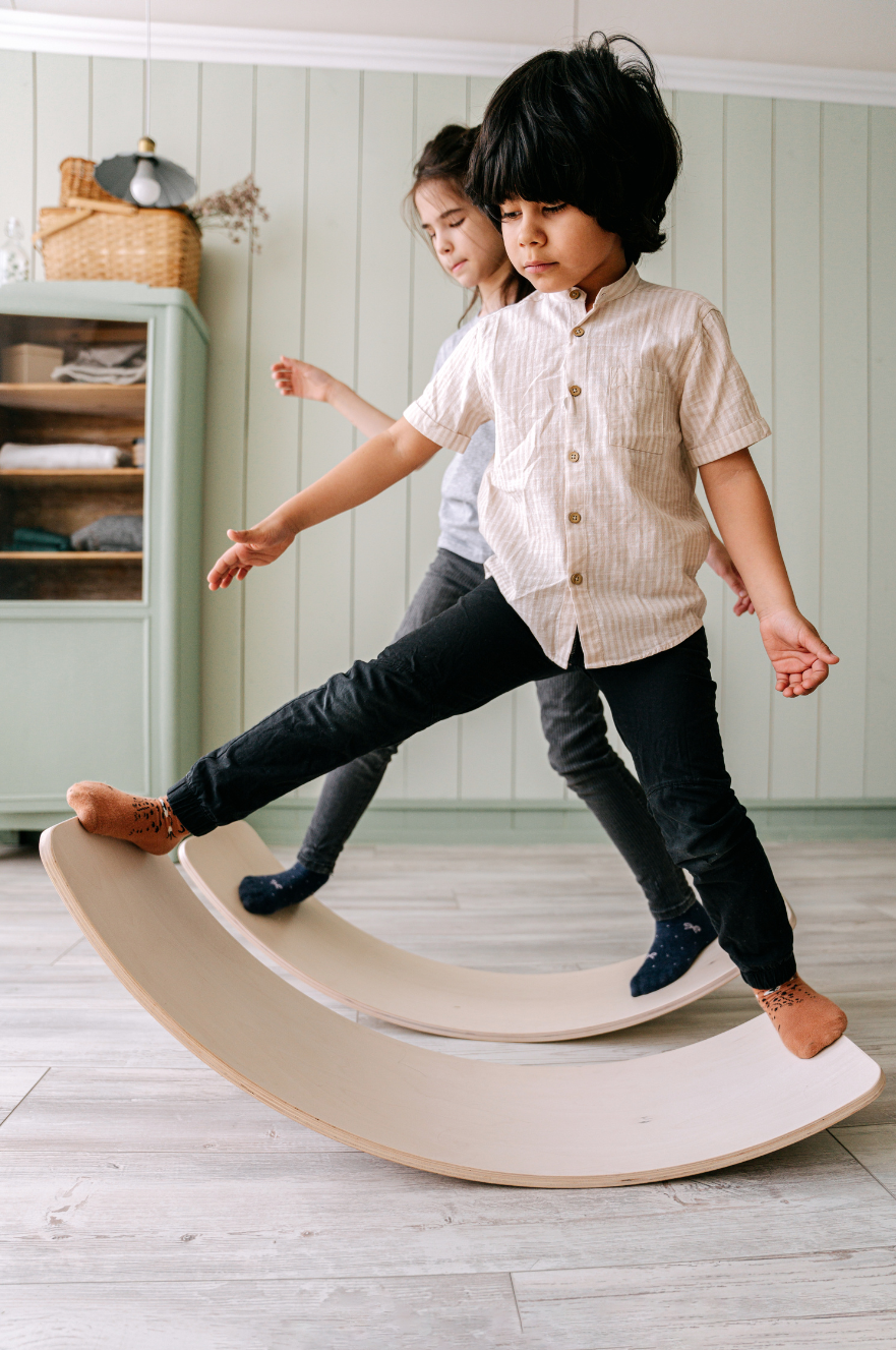 Two children balancing on a curved wooden board indoors, with one boy in a beige shirt and black pants and a girl in a gray shirt and dark pants.