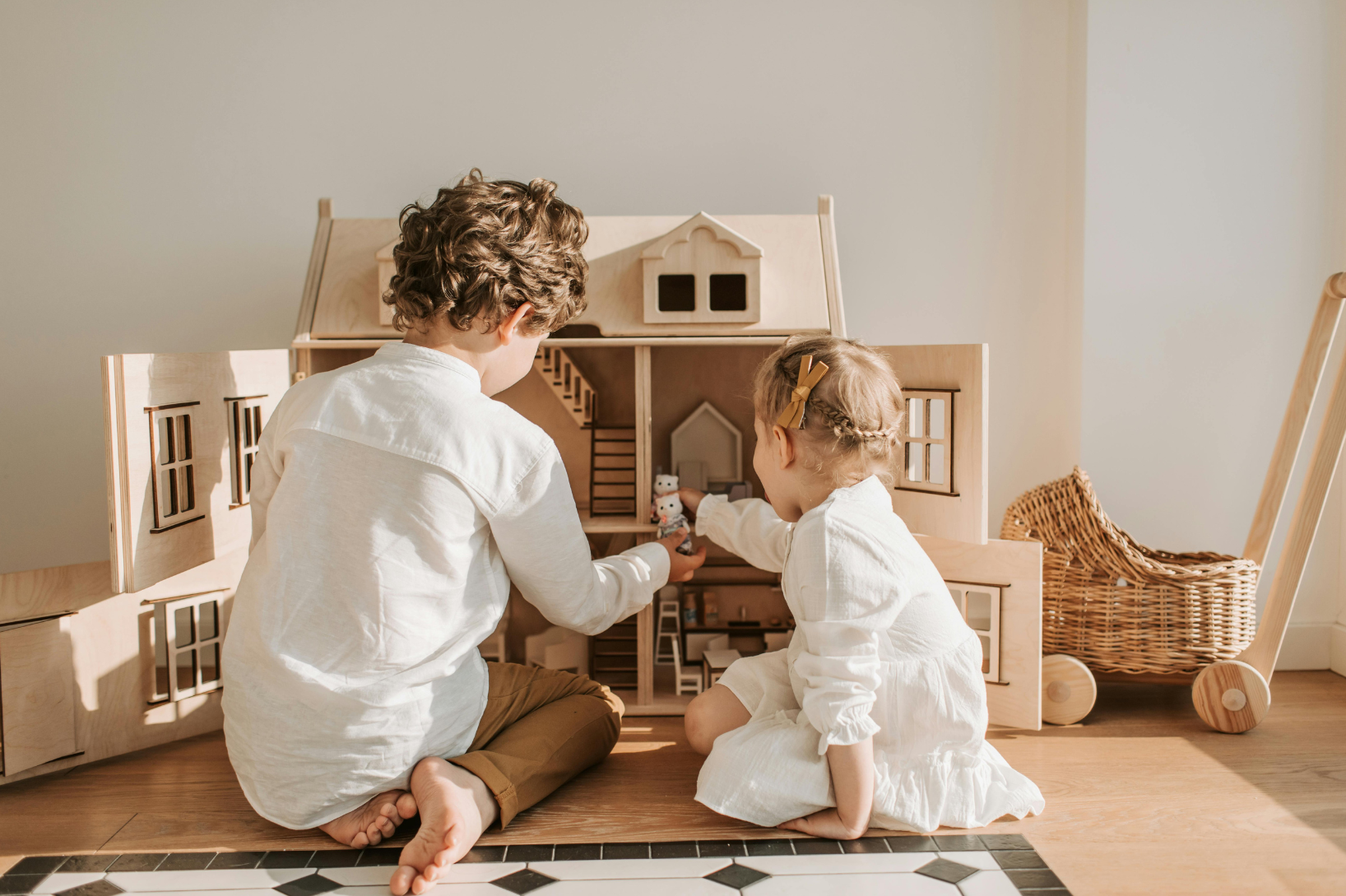 Two children, a boy and a girl, playing with a wooden dollhouse indoors. They are sitting on the floor, with the boy on his knees and the girl sitting cross-legged, both dressed in white.
