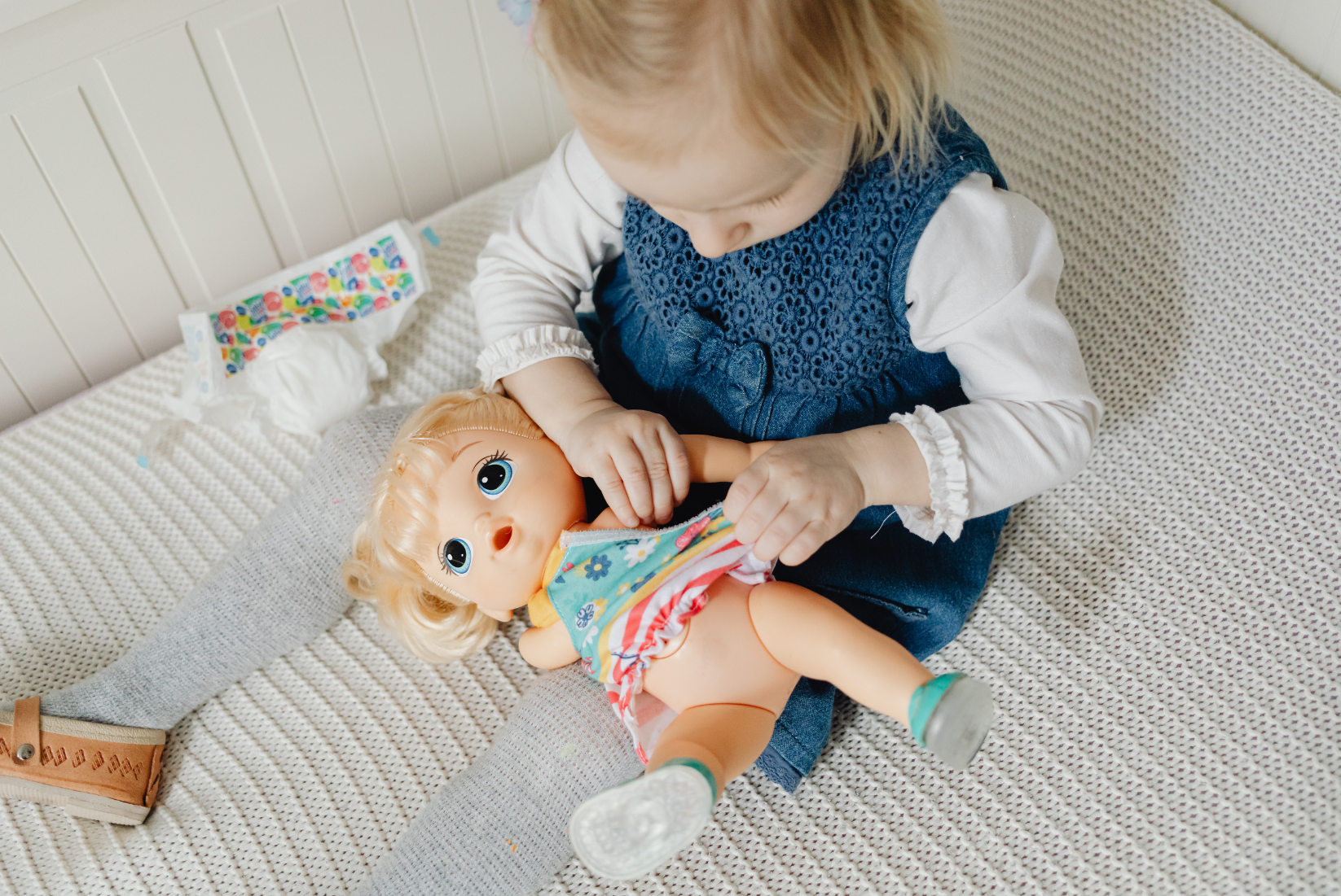 A young girl in a blue dress and gray tights is playing with a doll on a beige textured surface.