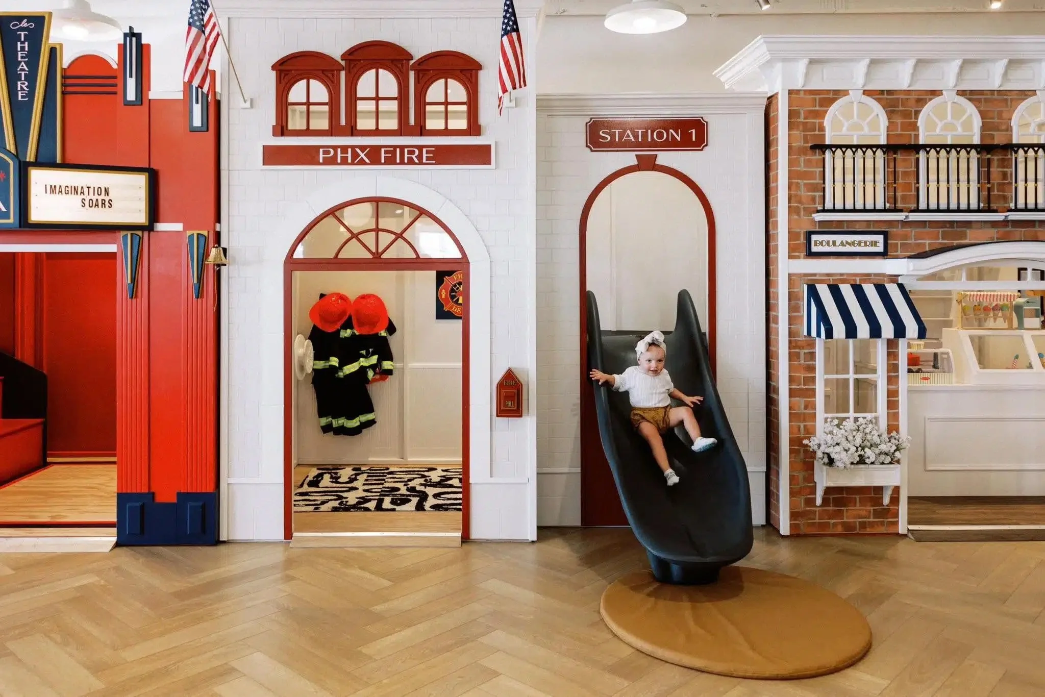 Child on a slide in an indoor play area designed to look like a fire station, with firefighter gear on the wall and signs reading 'PHX FIRE' and 'STATION 1'.