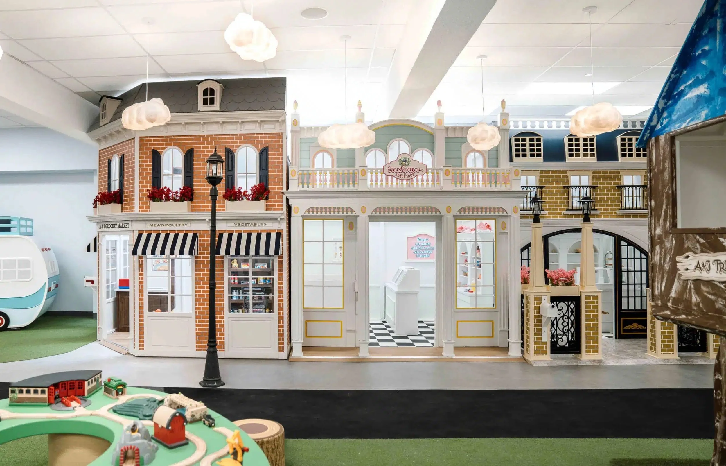 A toy or model village with shops, including a bakery and a boutique, with decorative clouds hanging from the ceiling and a miniature train and track in the foreground.