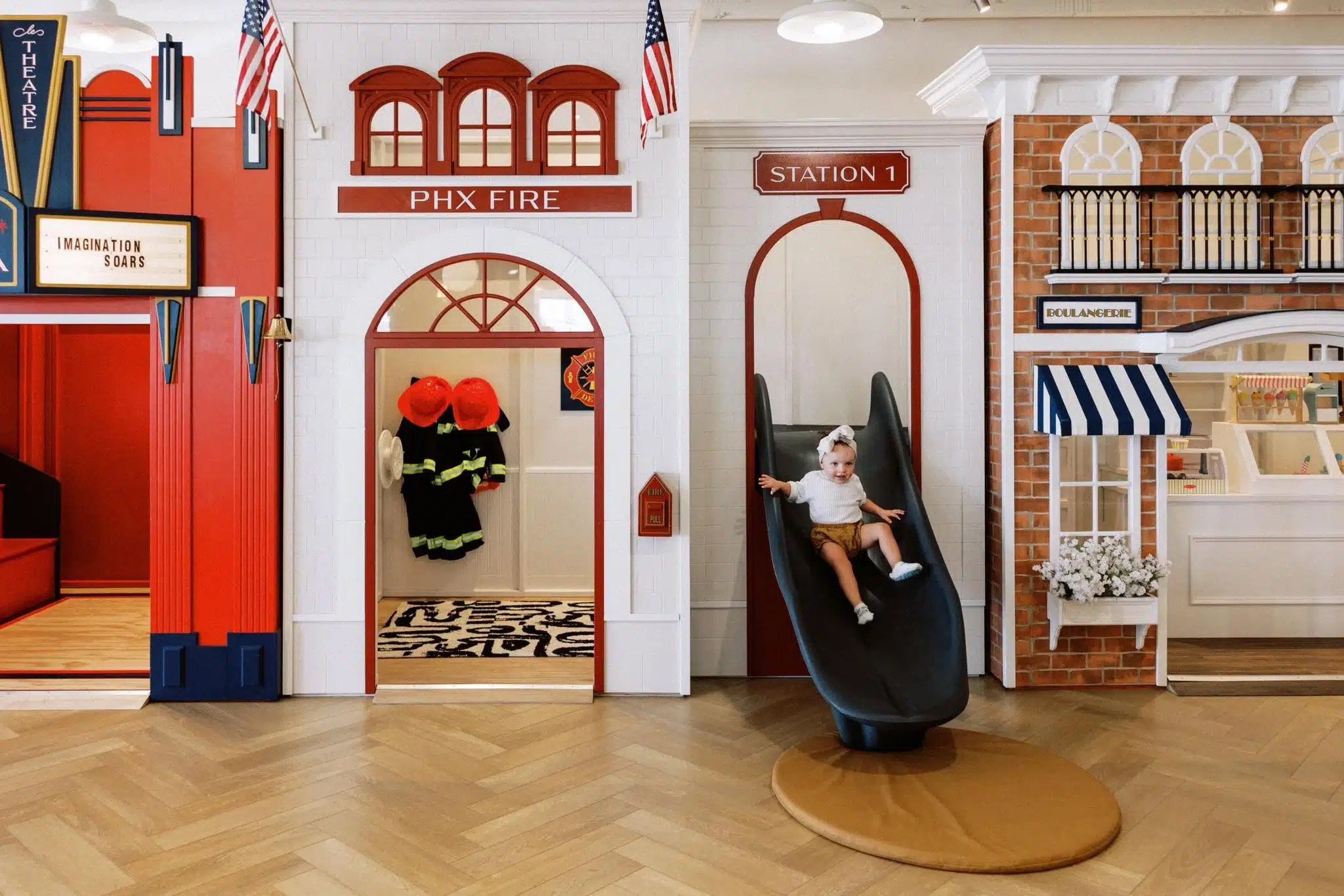 Interior of a children's play area designed like a fire station and bakery, featuring a small slide, fireman's coat and helmets, signage for 'PHX FIRE' and 'STATION 1', and bakery signage 'BOULANGERIE', with a young girl playing on the slide.