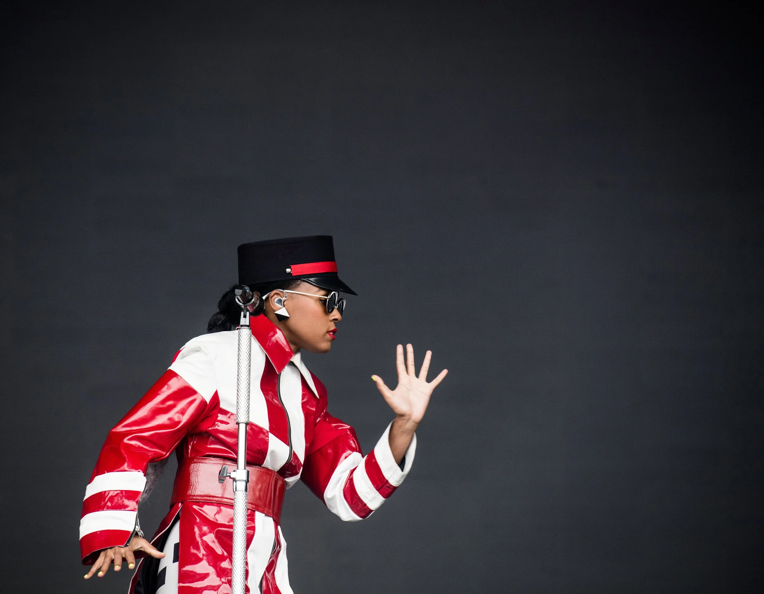 Woman in a futuristic red and white outfit with a black hat, sunglasses, and wireless earbuds, gesturing with her right hand against a dark gray background.