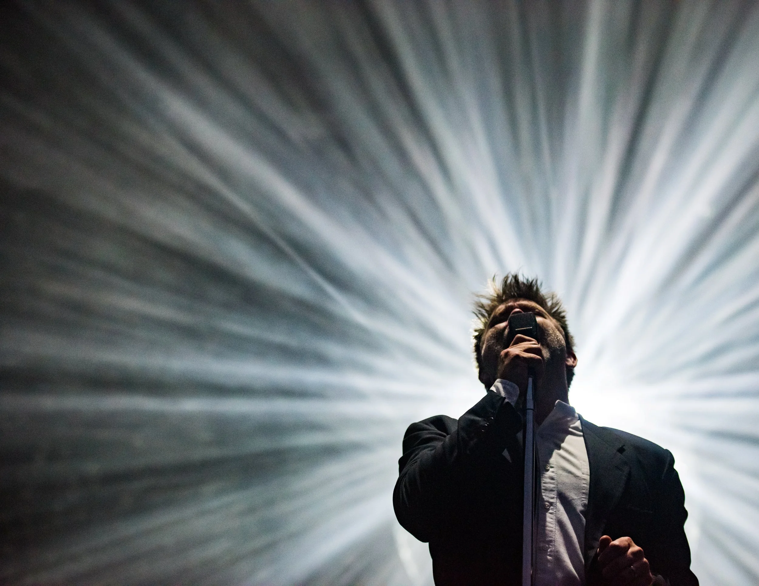 A man singing into a microphone on stage with dramatic lighting from behind, creating radiating light effects.