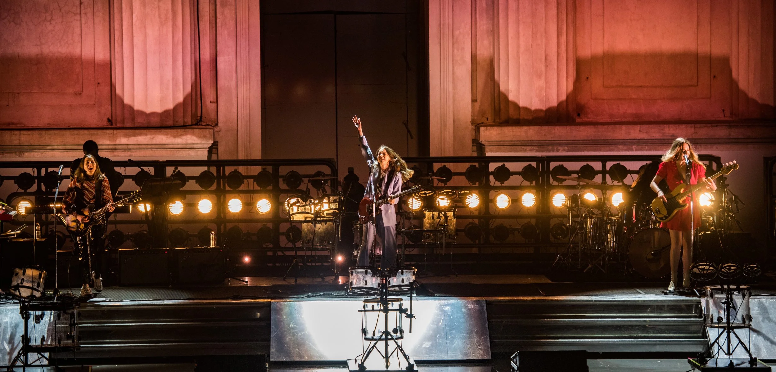 A band performing on stage with musicians playing guitars, drums, and bass, with stage lights and a large wooden backdrop.