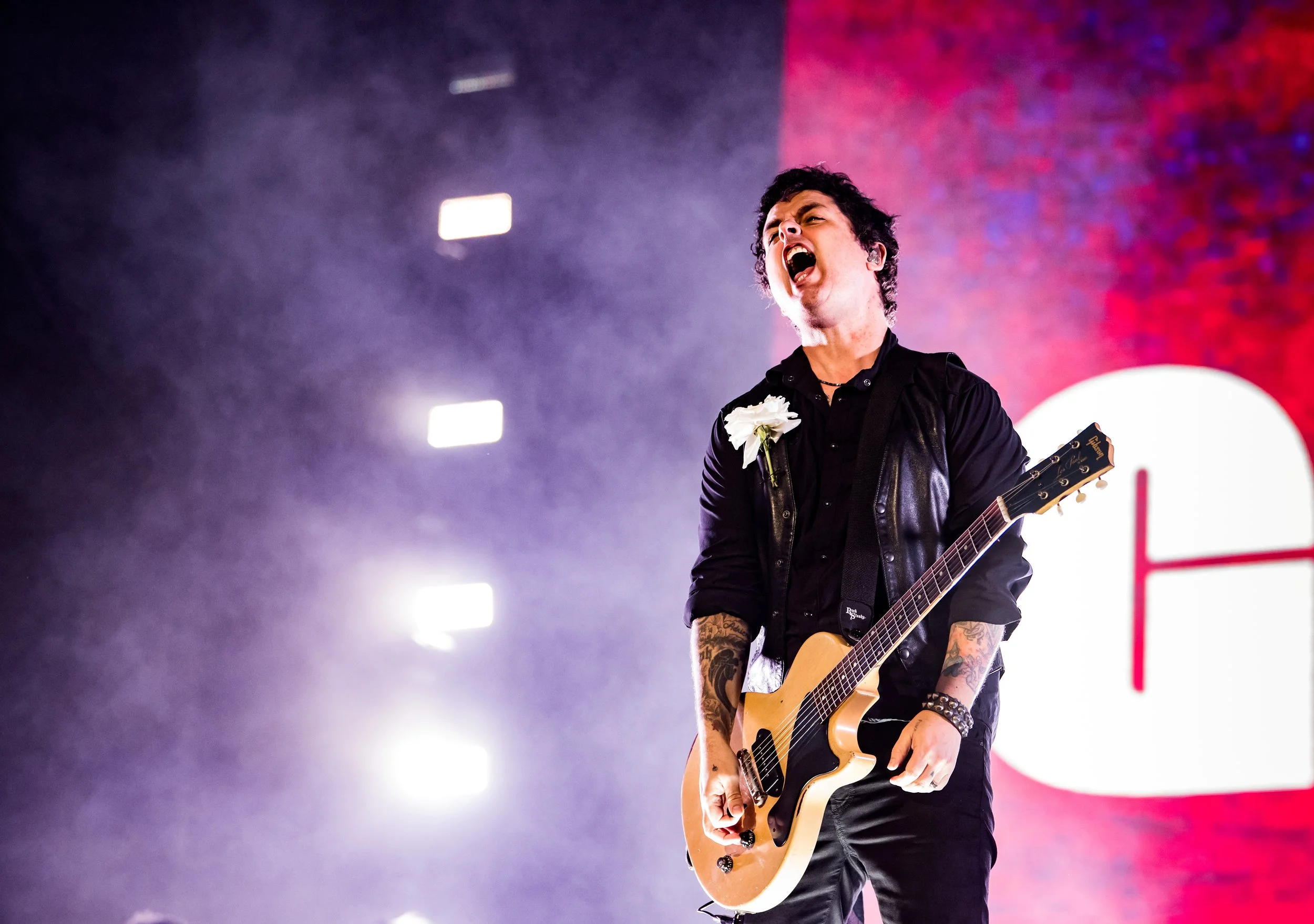 A male guitarist performing on stage, singing passionately with his mouth open, wearing a black shirt and leather vest, with tattoos on his arms, holding an electric guitar, against a background of bright stage lights and a red-lit wall.