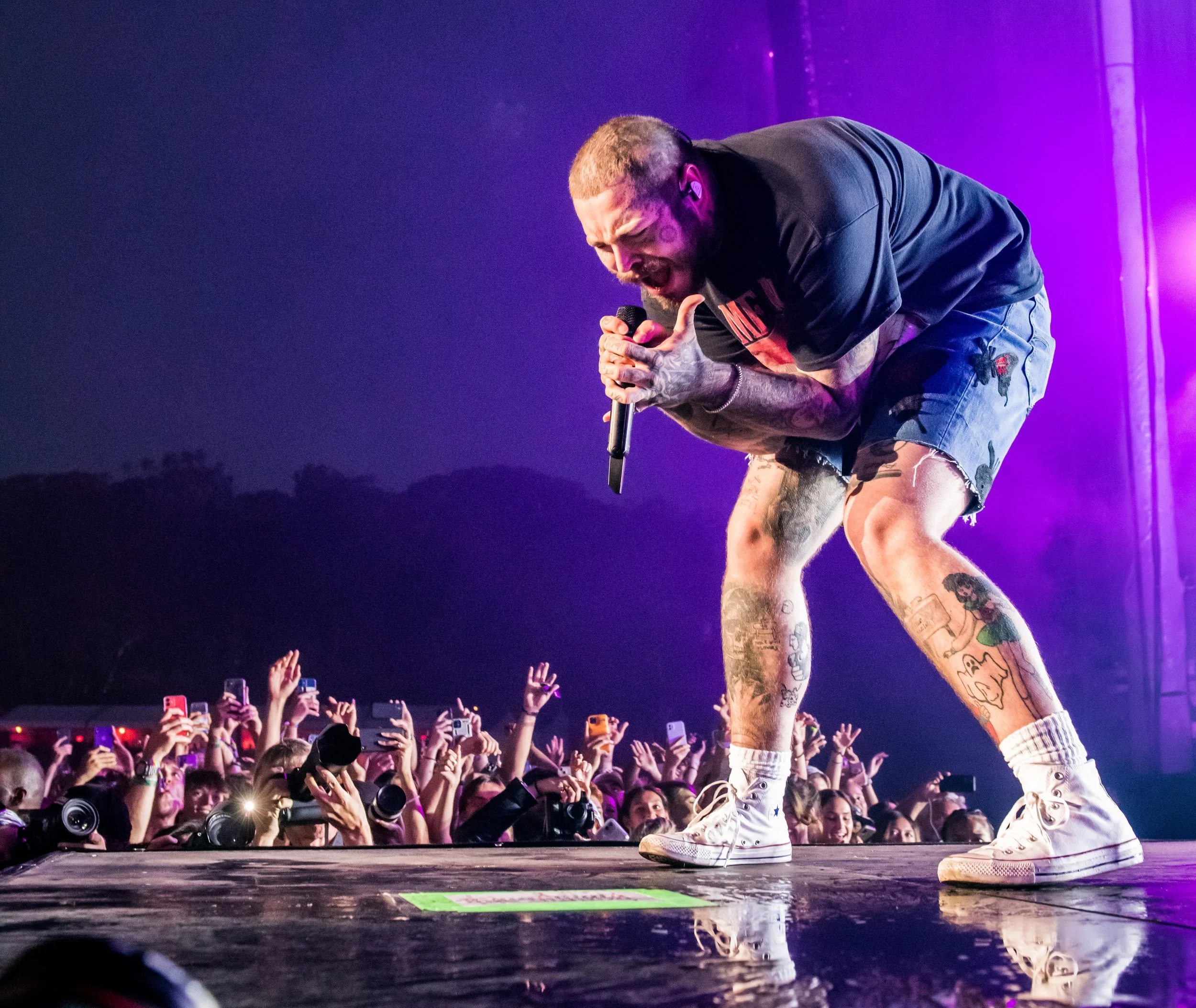 Male singer performing passionately on stage with crowd taking photos and raising hands in the background, purple stage lights.