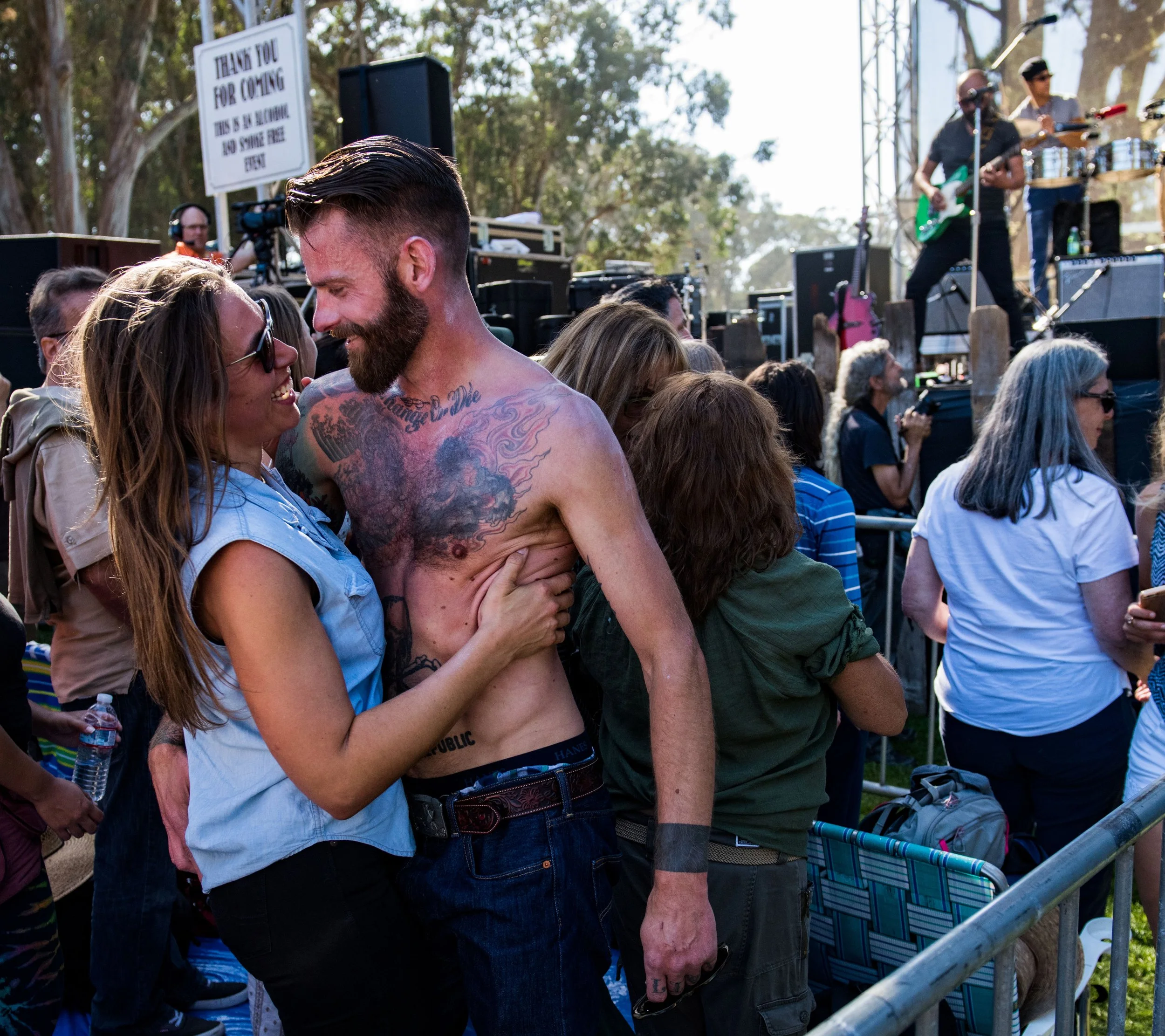 A couple at an outdoor concert, with the woman wearing sunglasses and a sleeveless shirt, and the shirtless man with tattoos, smiling and looking into each other's eyes. They are surrounded by other concert attendees, with a stage and band performing
