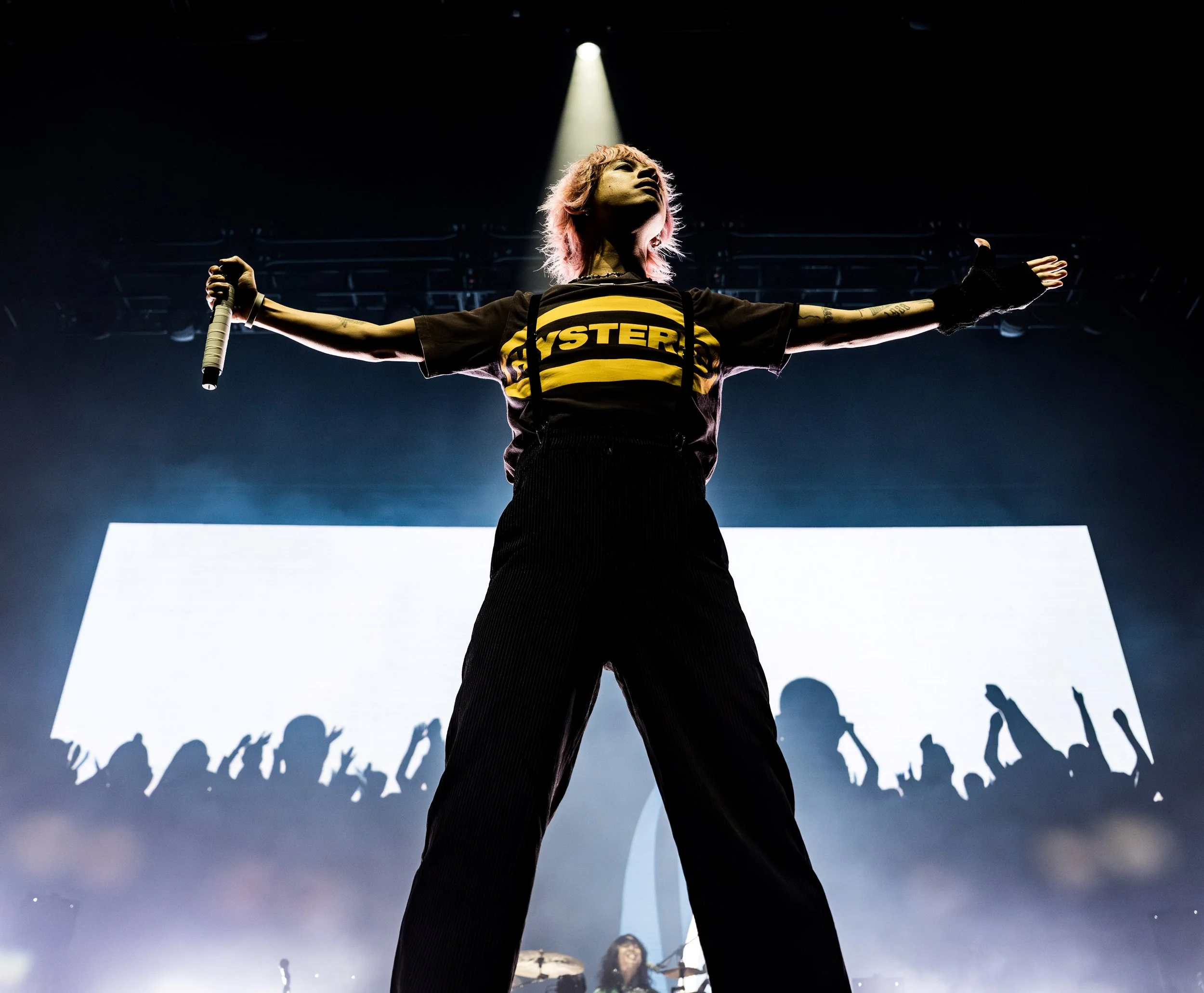 A female performer stands on stage with arms outstretched, holding a microphone in her right hand, under a spotlight. She is wearing a black and yellow shirt with the word 'HYSTERIA' on it and black pants, with a crowd silhouette visible in the backg