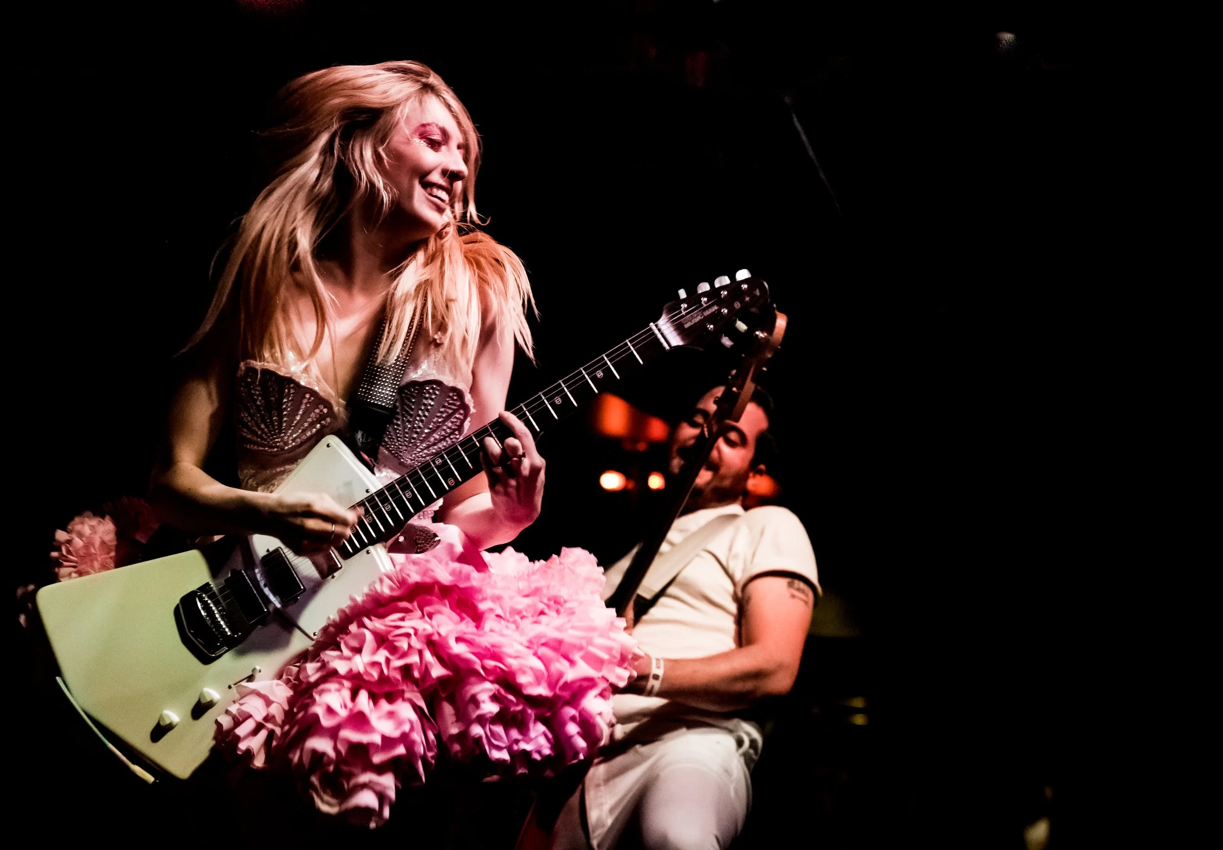A woman with blonde hair playing an electric guitar, smiling, during a performance with a male musician also playing guitar in the background, in a dimly lit setting.