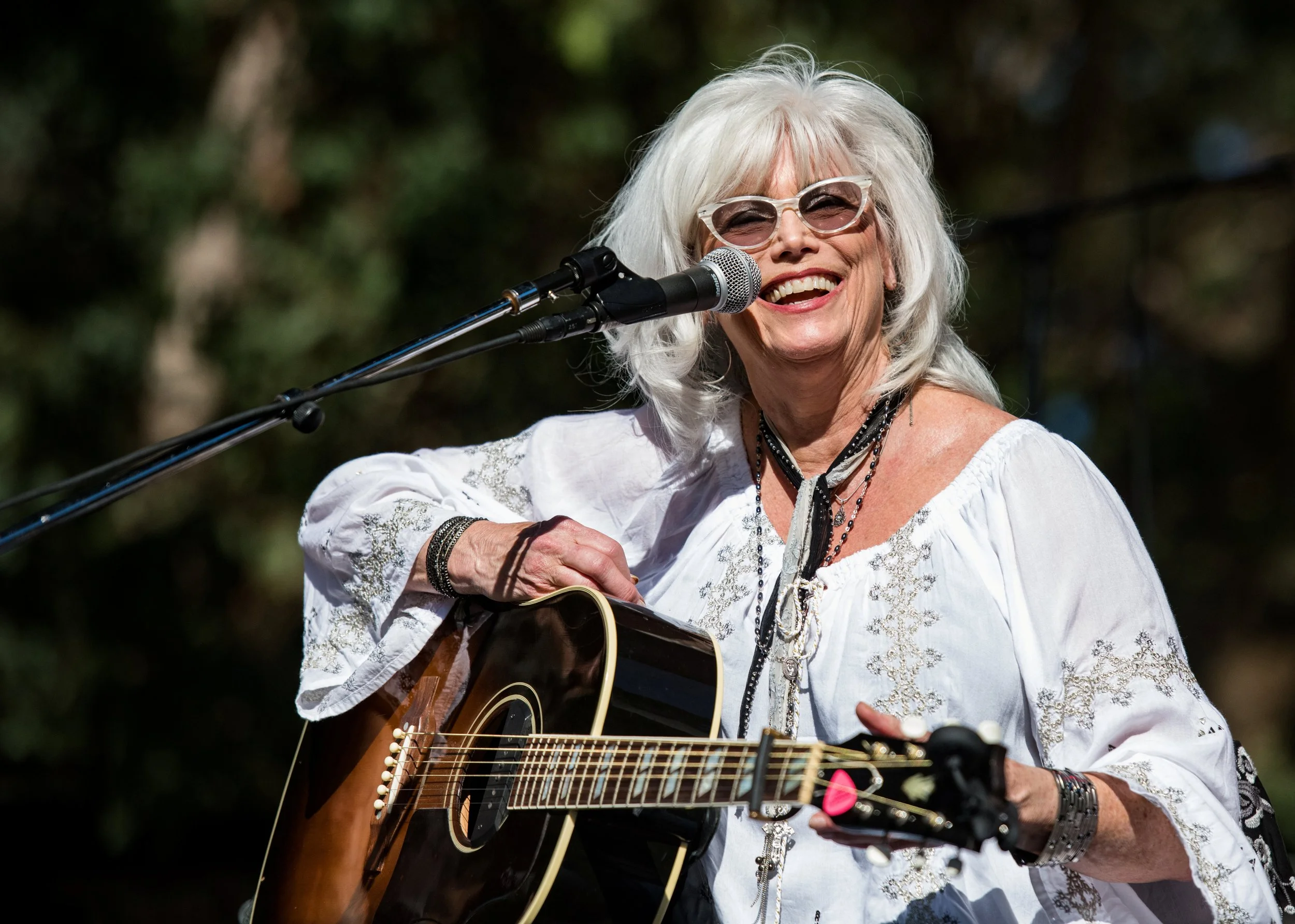 An elderly woman with white hair and glasses playing an acoustic guitar and singing into a microphone outdoors.