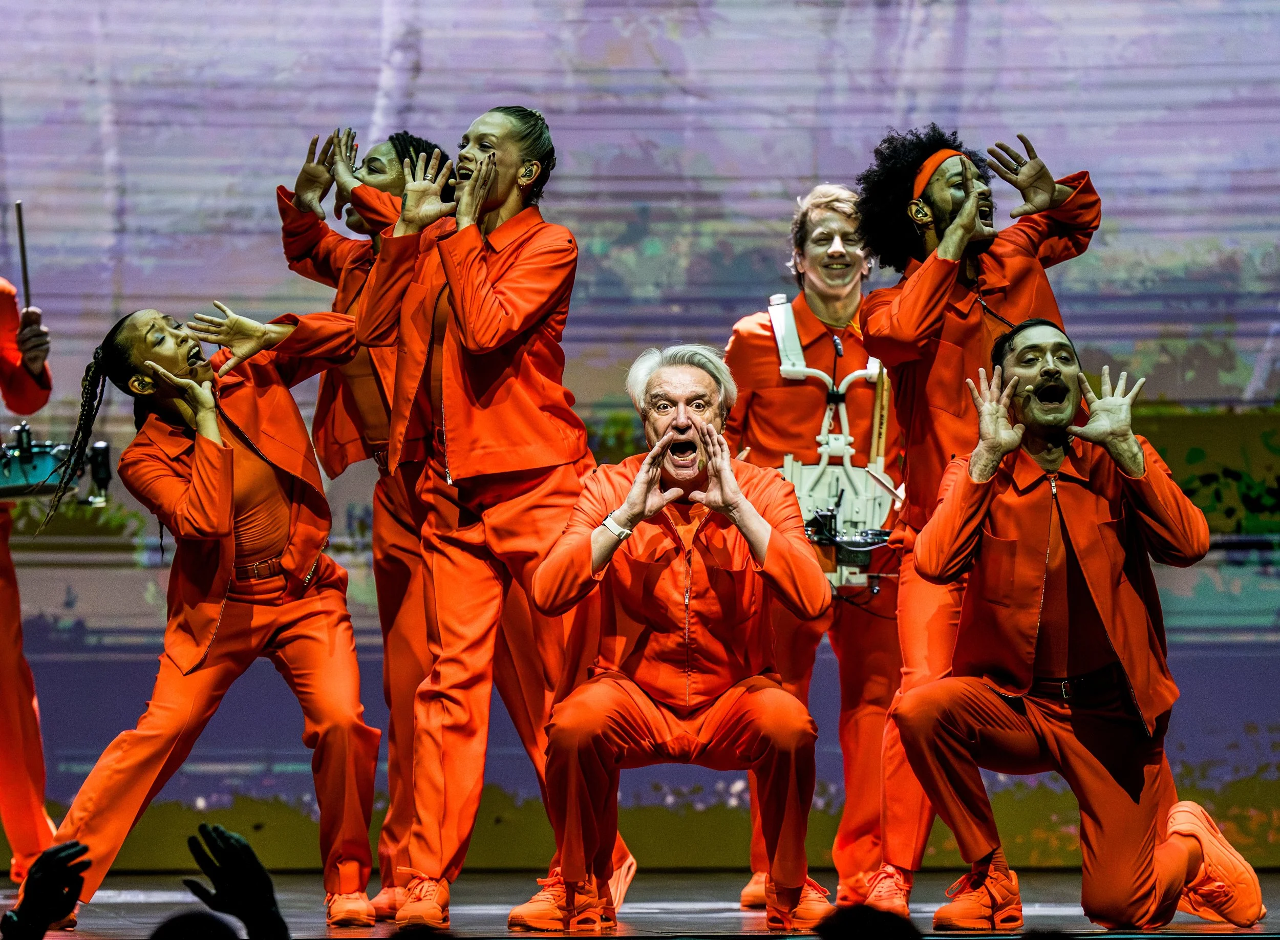 Group of performers dressed in orange jumpsuits on stage, making energetic and expressive gestures, with colorful background and audience in foreground.