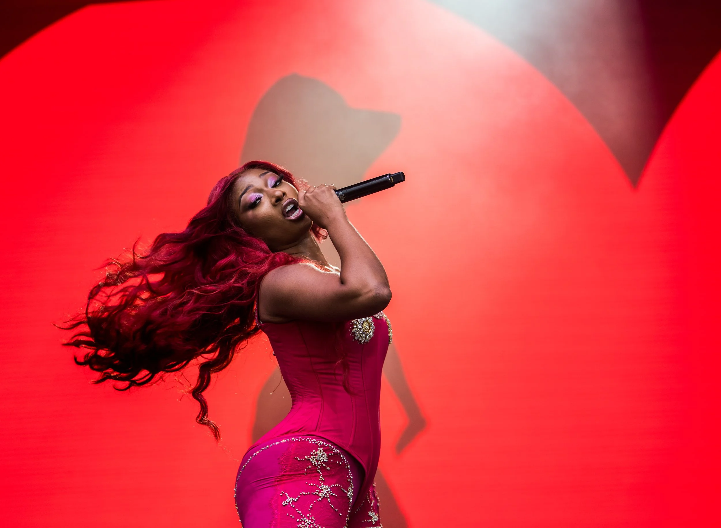 A woman with long red hair singing into a microphone on stage with a red background.