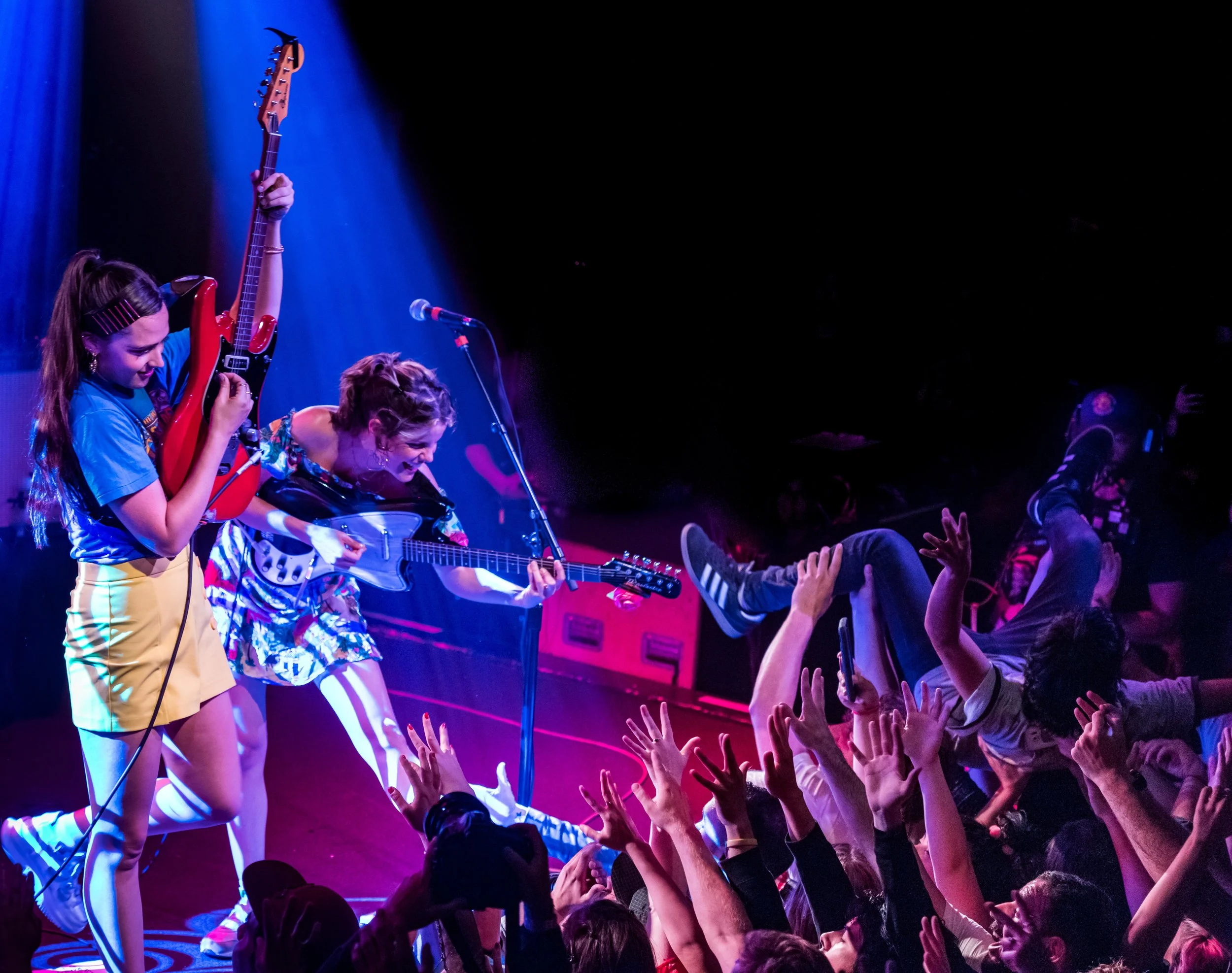 Two young women performing on stage with guitars while a person crowd surfs, reaching out with hands to touch them, during a concert.