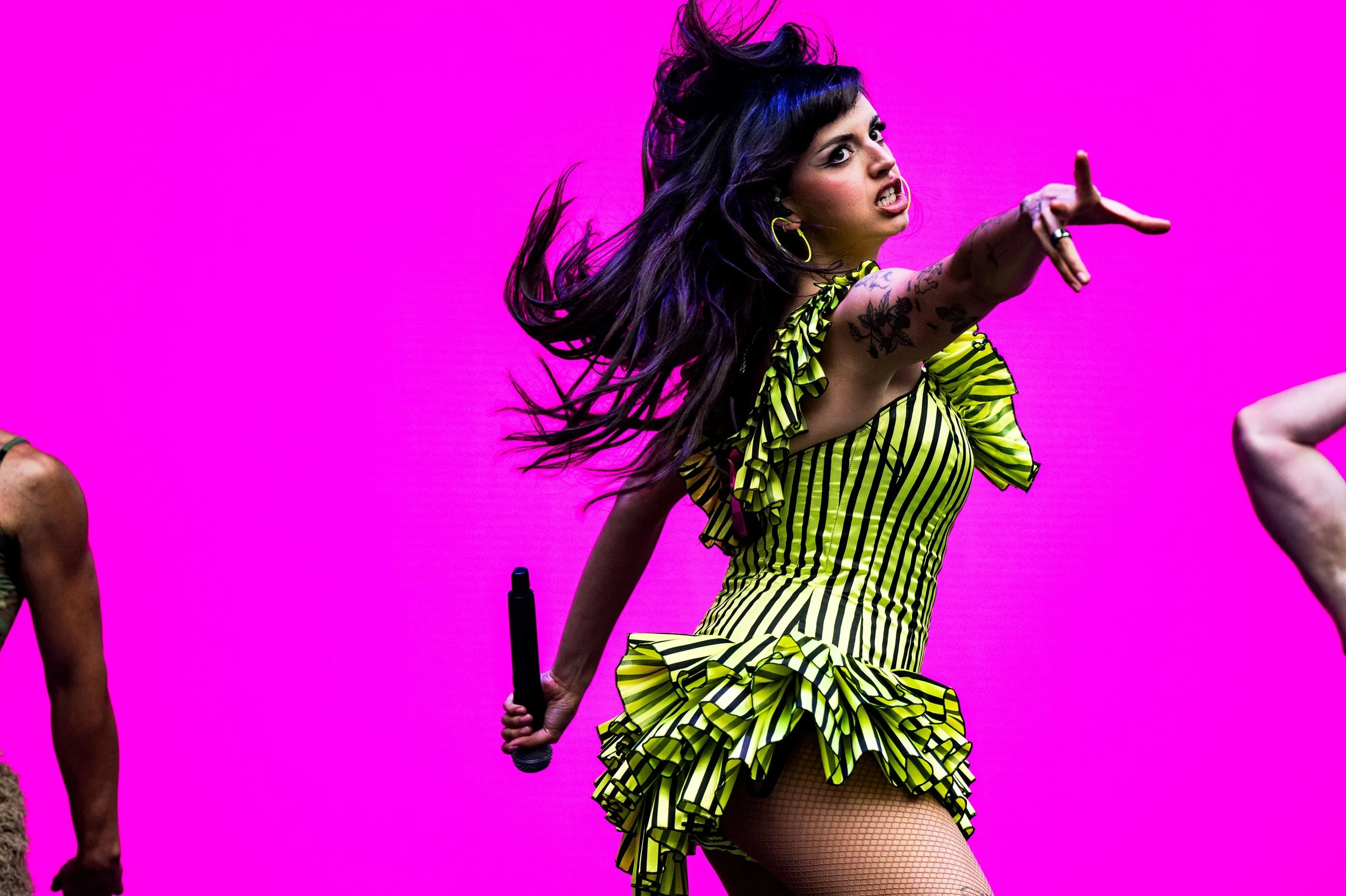 A woman with long dark hair, tattoos, and yellow earrings, wearing a yellow and black striped ruffled dress, is pointing with her right hand and holding a microphone in her left hand against a vibrant pink background.